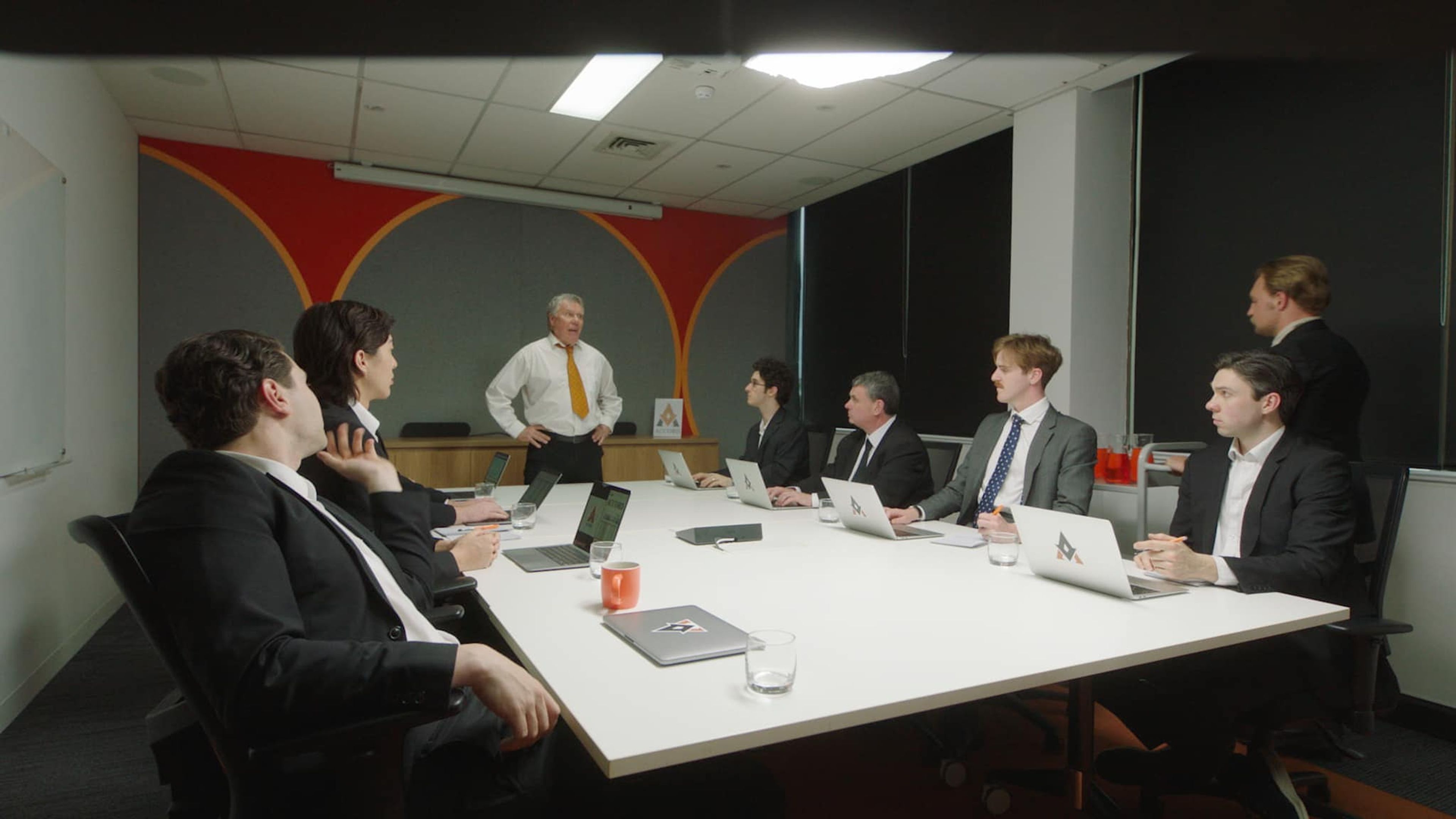 Film still of a business meeting in a boardroom with seven men in suits listening to an older man standing and speaking.