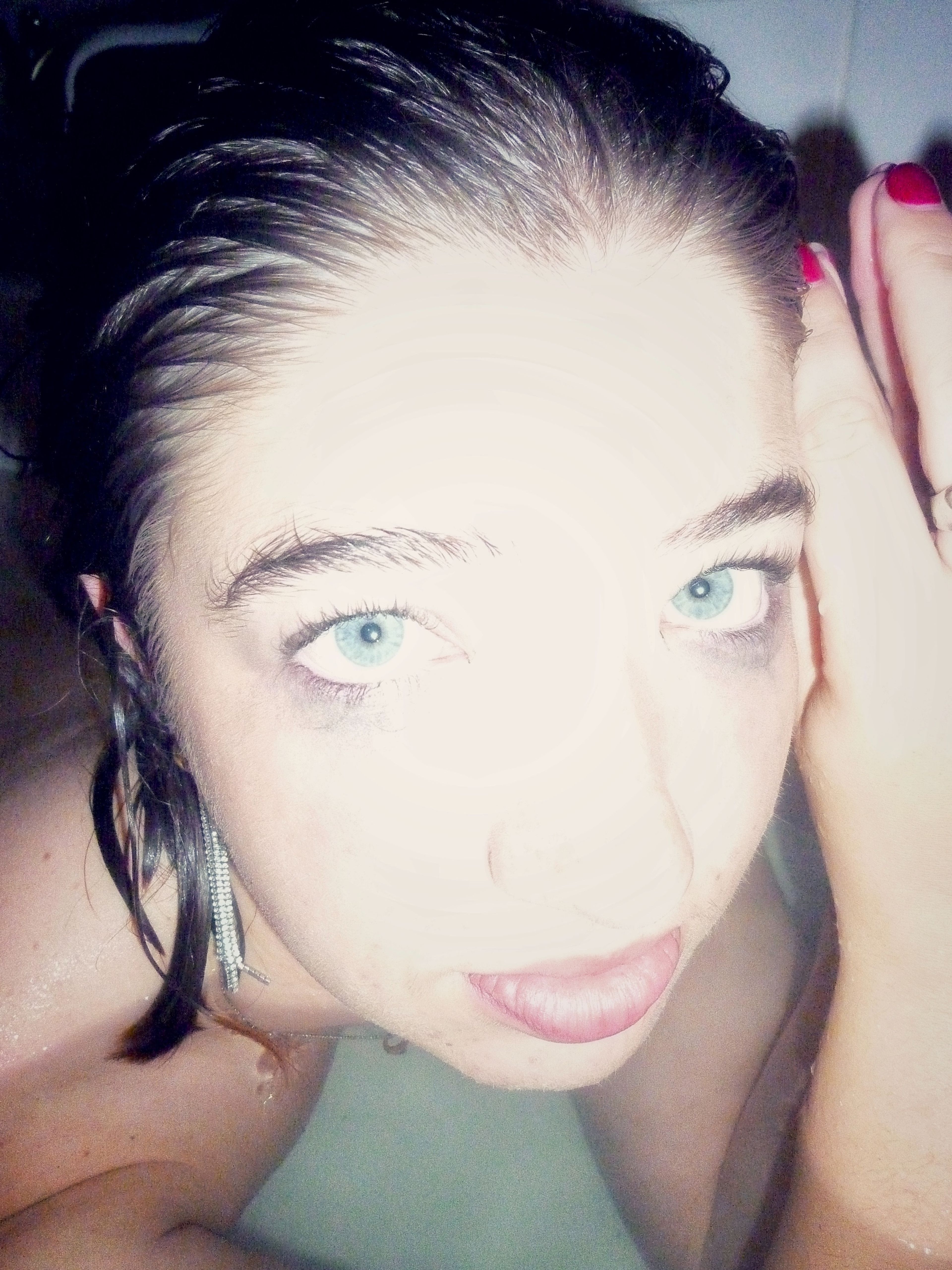 A stylized portrait photograph of a woman in a close-up shot. She is leaning in a bathtub, with bright lighting emphasizing her intense blue eyes and facial features, contrasting against her dark, wet hair and slightly smudged makeup.