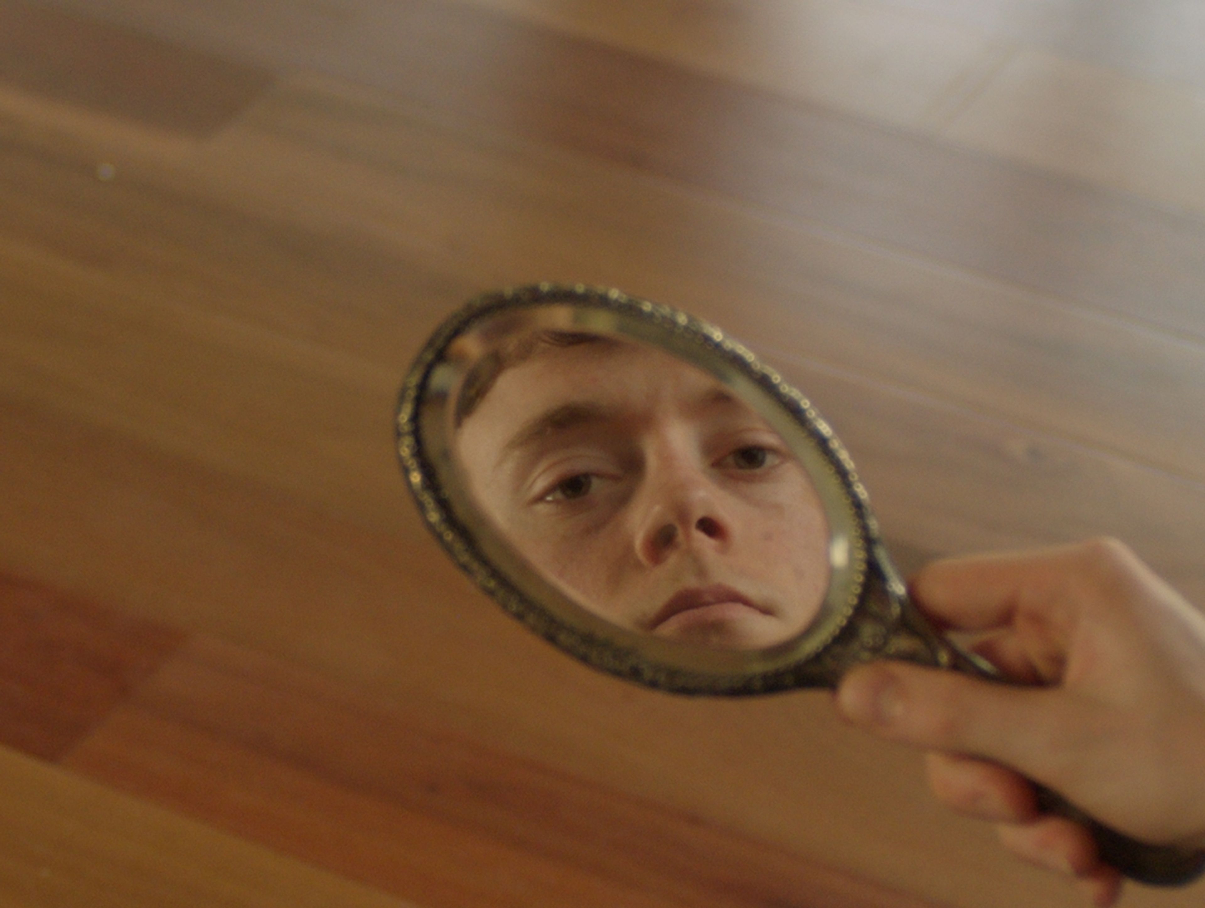 Film still showing a close-up reflection of a young person's solemn face in an ornate hand mirror against a wooden background, shot with warm lighting.