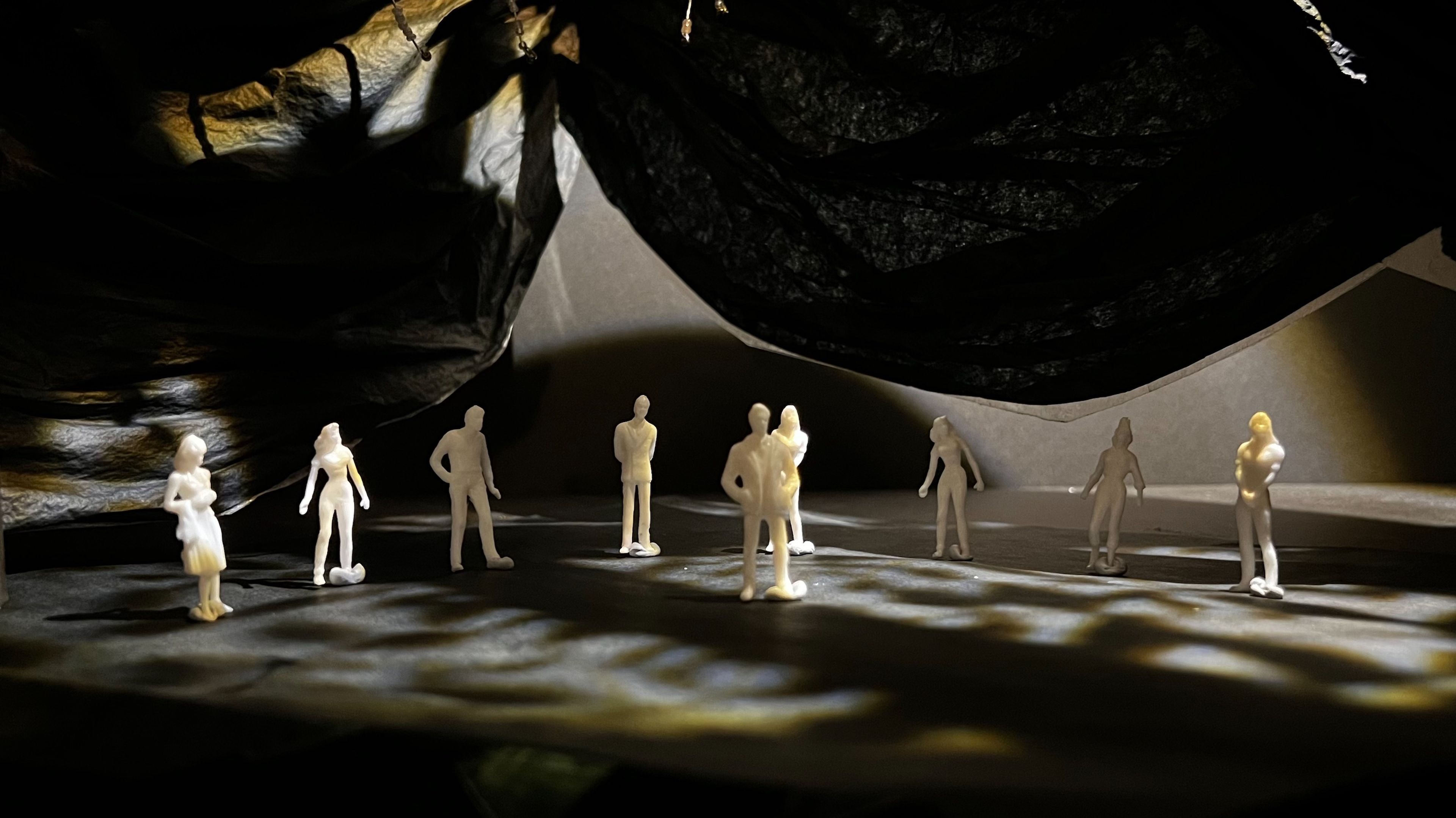 Scale model of a theatrical set showing eight white figures arranged in a semi-circle on a dark stage, with textured black fabric creating a cave-like ceiling