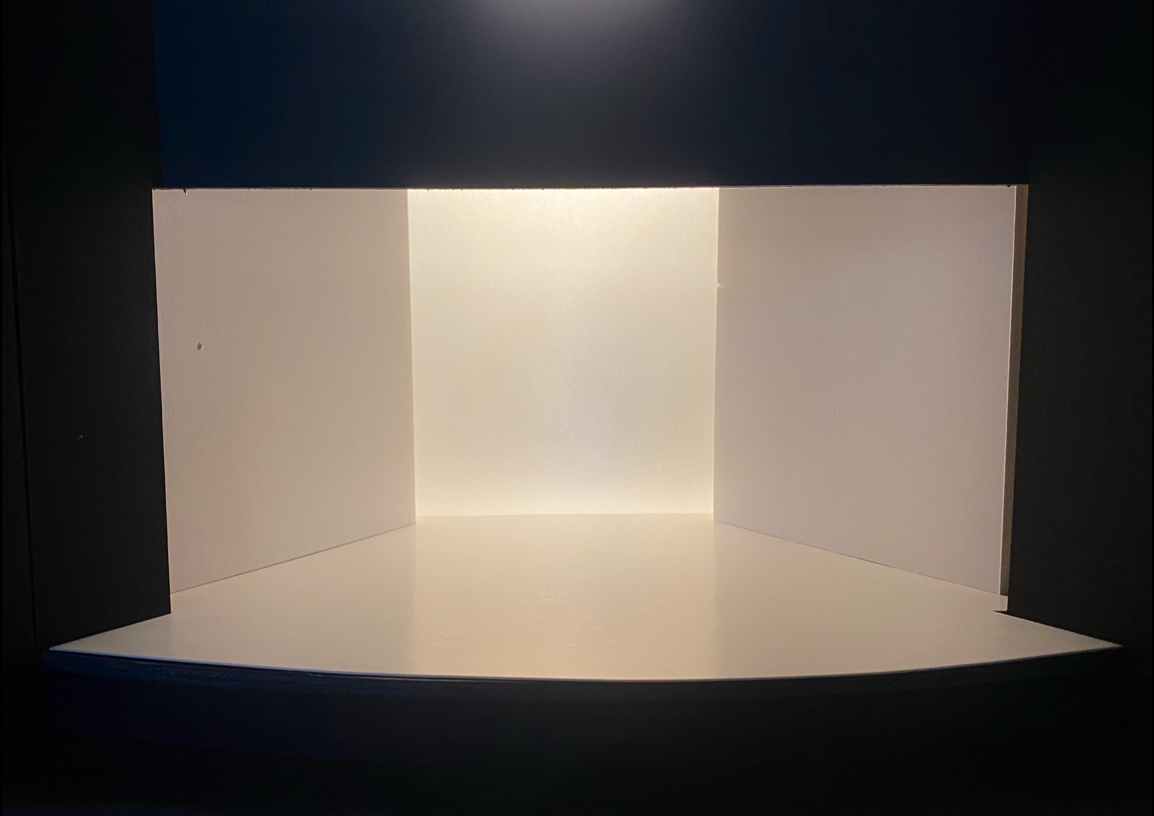 A minimal set model with white walls and a white floor, softly illuminated. The empty space provides a blank canvas for potential staging.