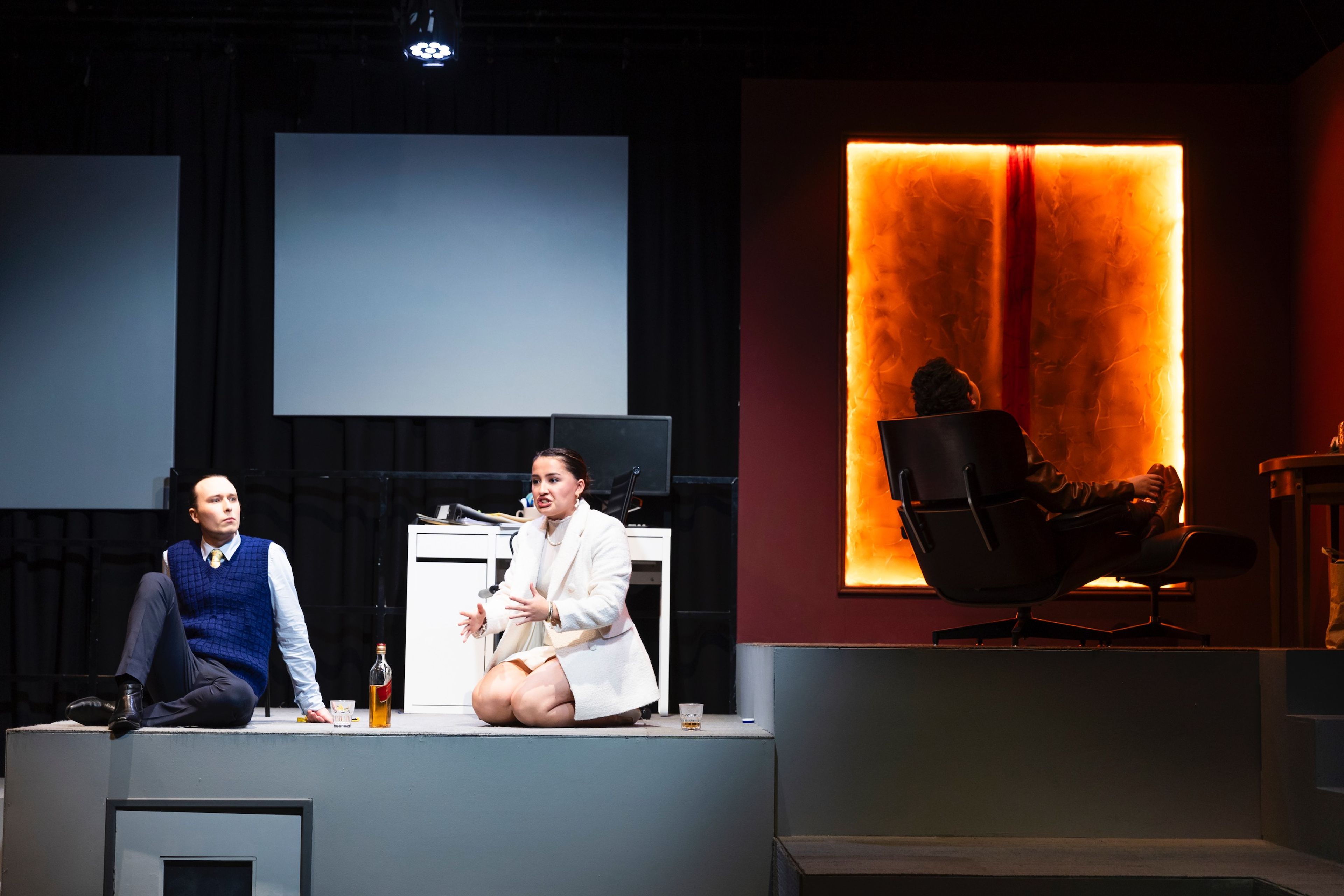 Theatre production photograph showing performers on a multi-level set - two seated on a grey platform with a bottle between them, and a silhouetted figure in a chair against an illuminated orange panel.