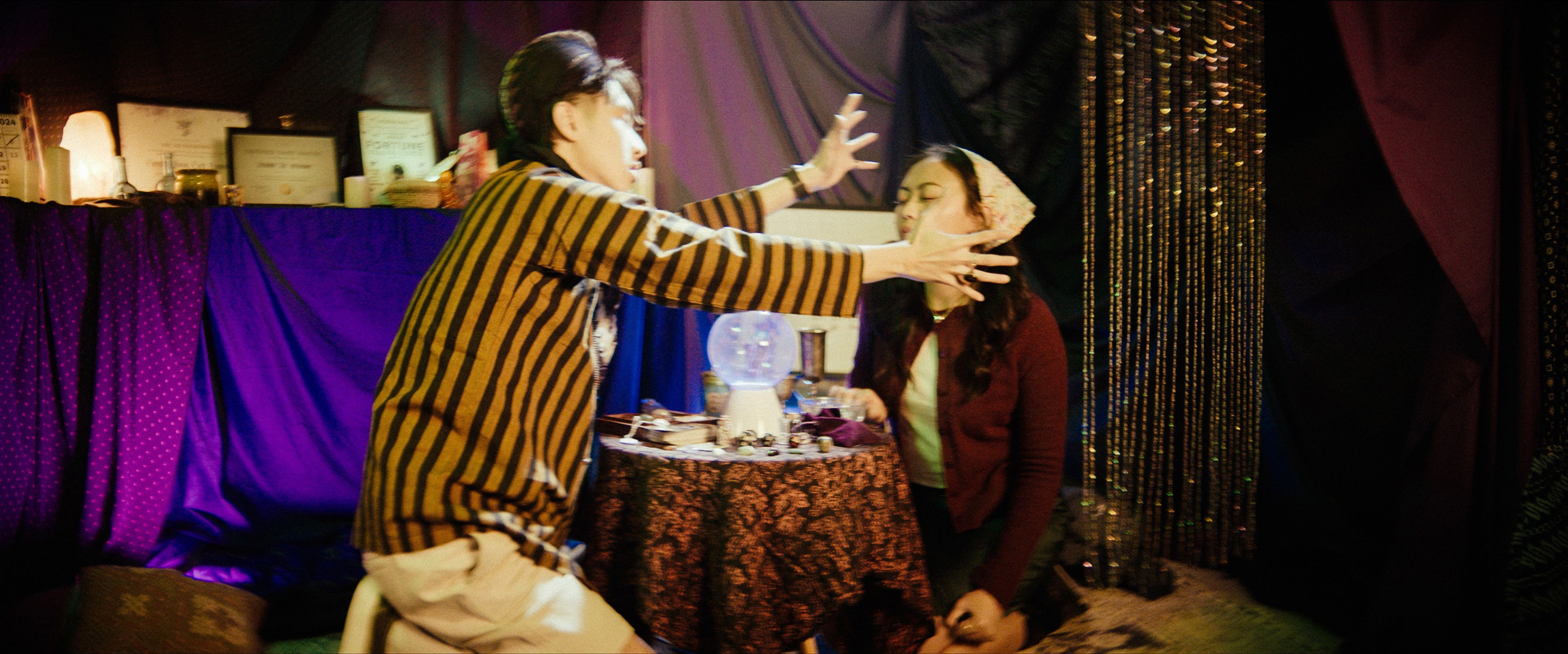 The image features two individuals in an animated interaction. On the left, a person wearing a yellow and black striped sweater extends their arms dramatically toward the person on the right, a crystal ball on the table between them