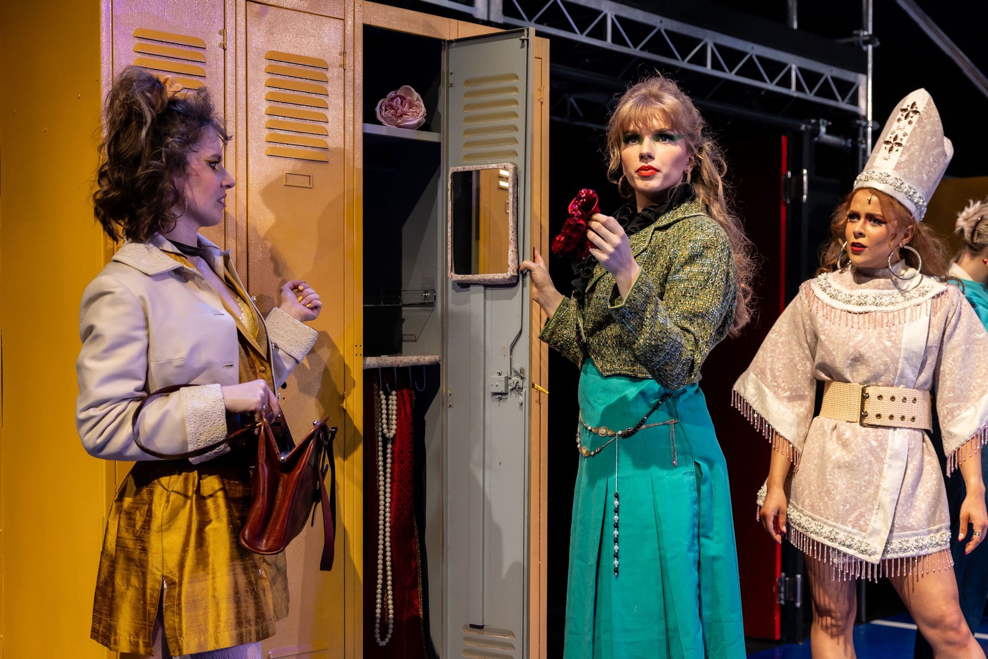 Theatre production photograph with three characters standing in front of lockers, dressed in colourful outfits, engaged in conversation.