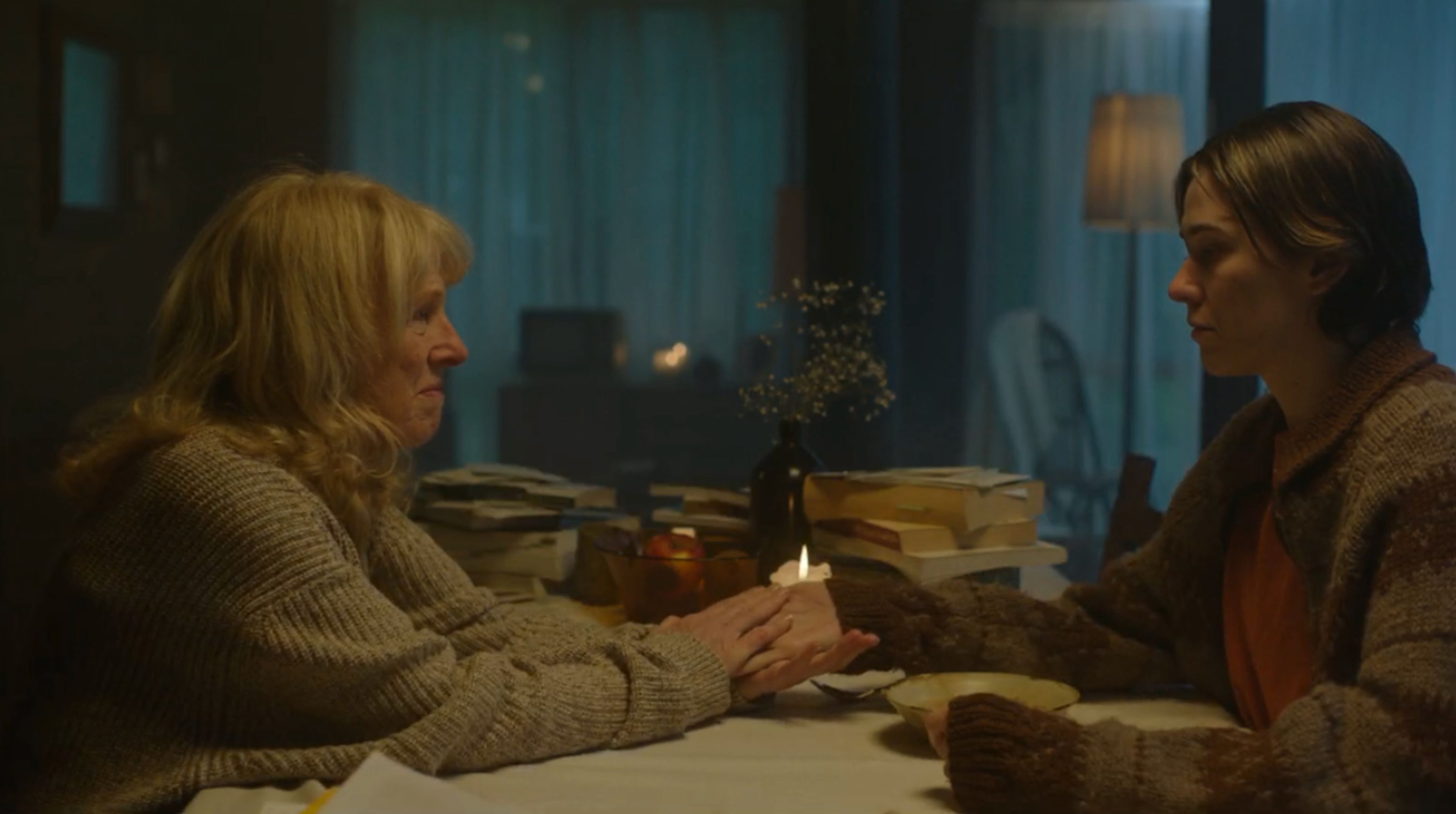 Film still depicting an intimate conversation between two women in chunky knit cardigans at a table, lit warmly by candlelight with stacks of books visible, creating a cosy atmosphere.