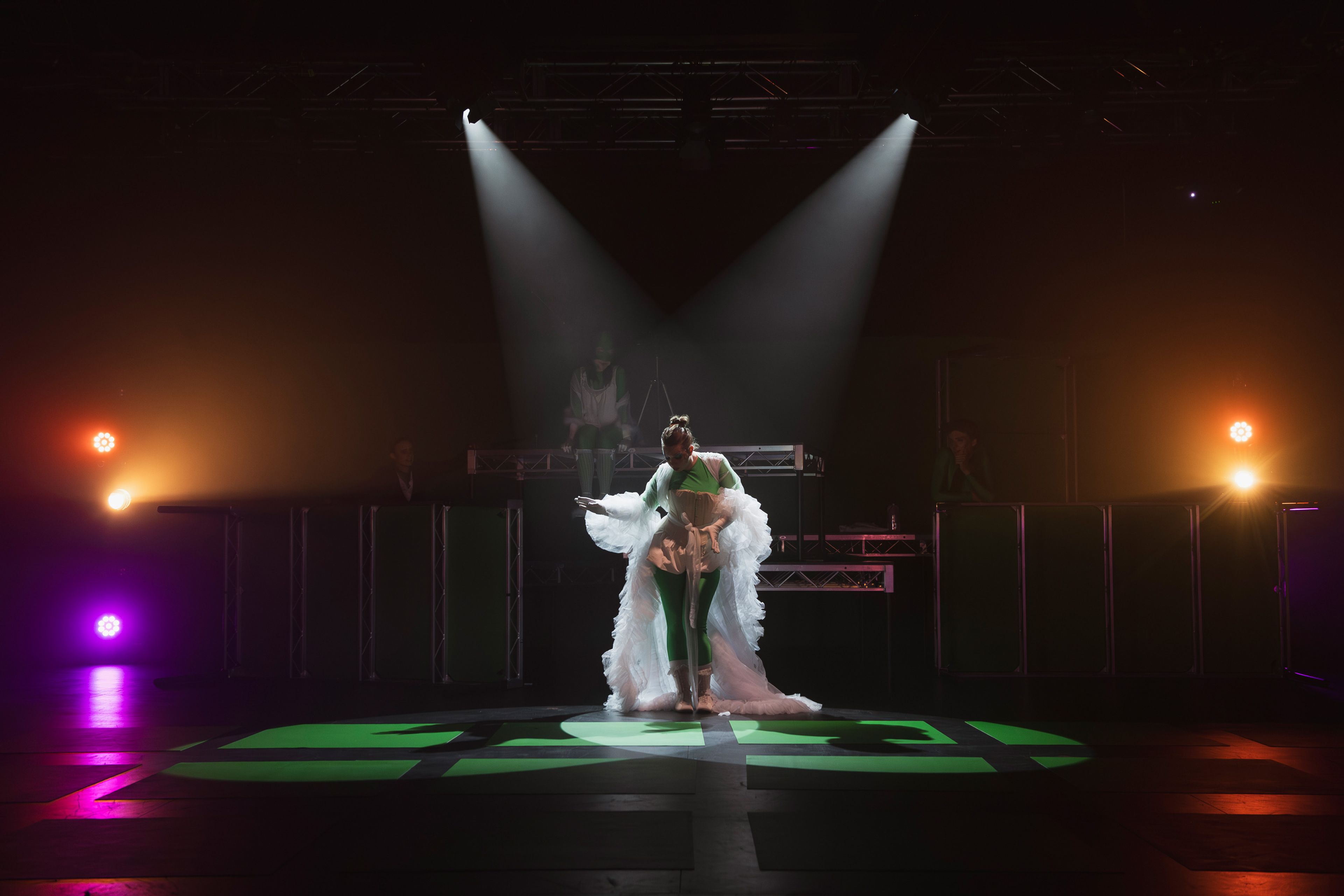 Performer in a flowing white costume illuminated by two spotlights, casting a striking silhouette. The background is dimly lit with vibrant orange and purple lights