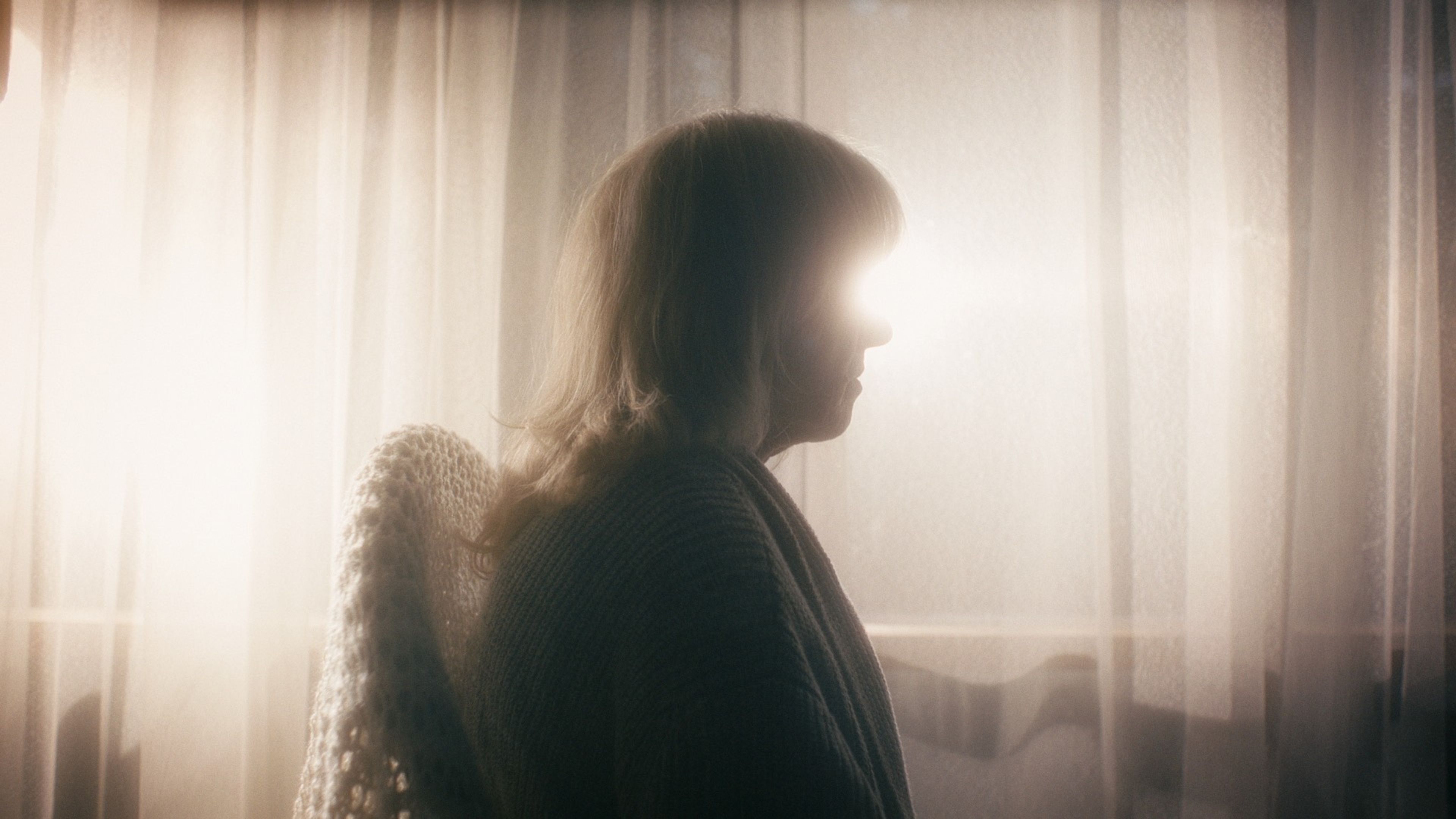 Film still featuring a woman's silhouette in profile against bright window light filtering through sheer curtains, wearing a dark cardigan.