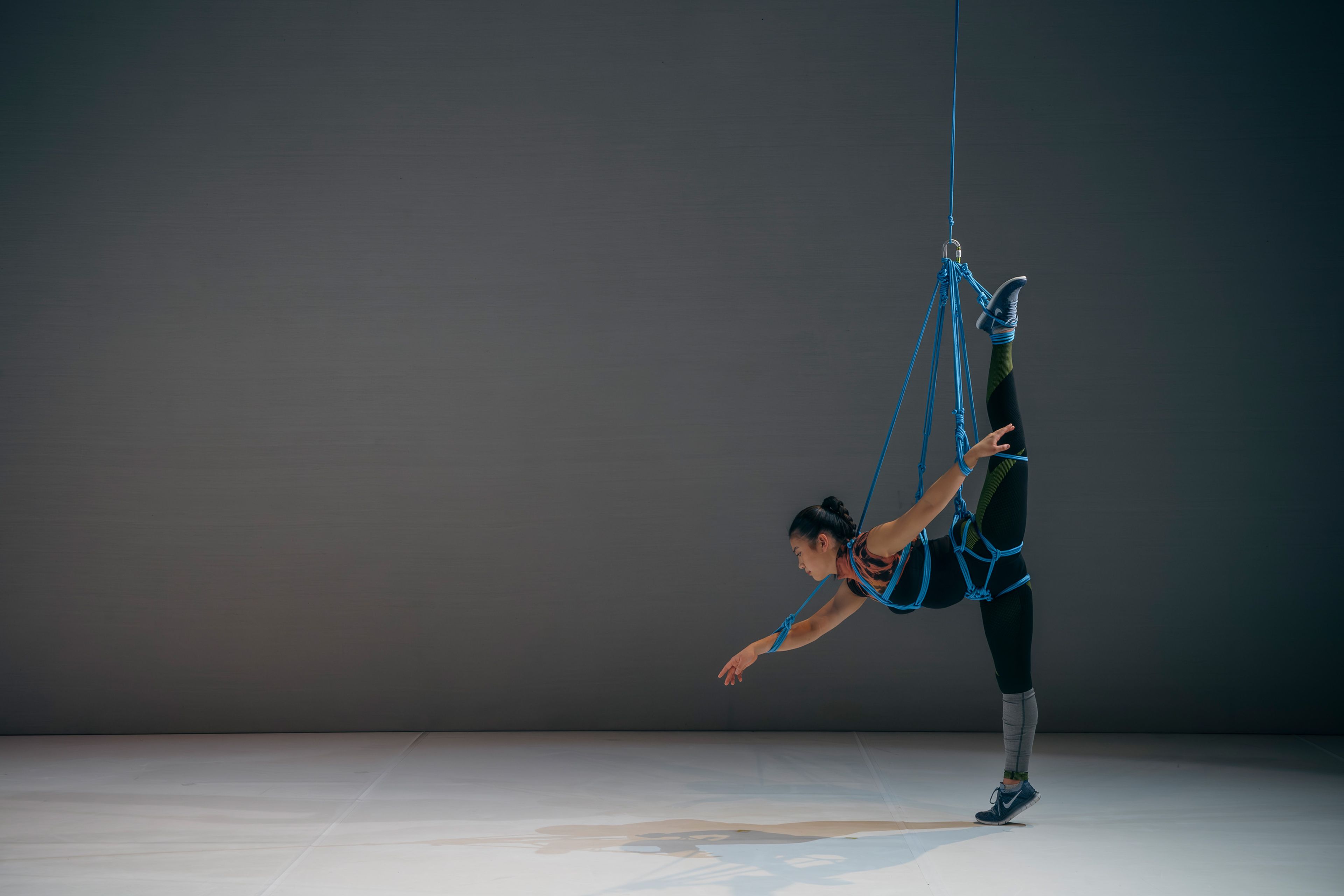 Dance production photograph of a performer in a blue aerial harness, stretching one leg upwards while suspended, with a minimalist grey background.