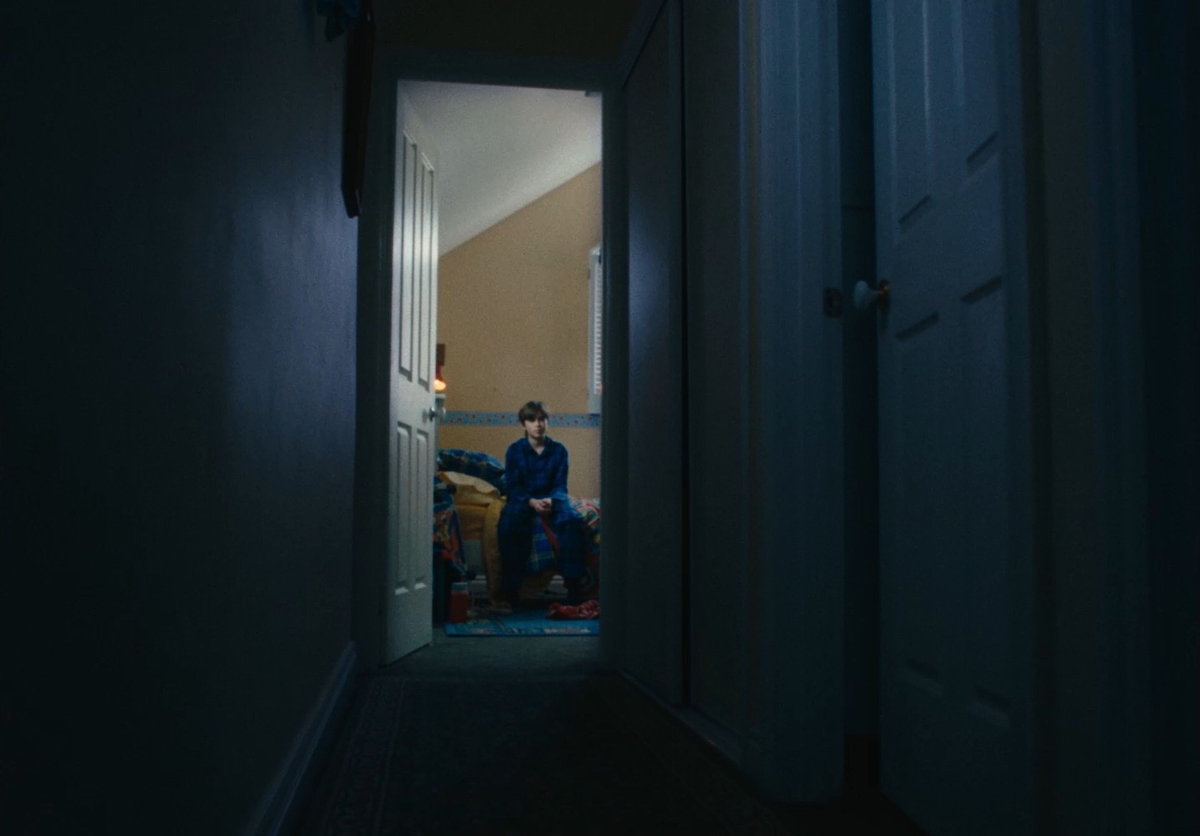 Film still looking down a dark hallway towards an open bedroom door, where a figure in blue clothing sits on the edge of a bed, lit from within the room.