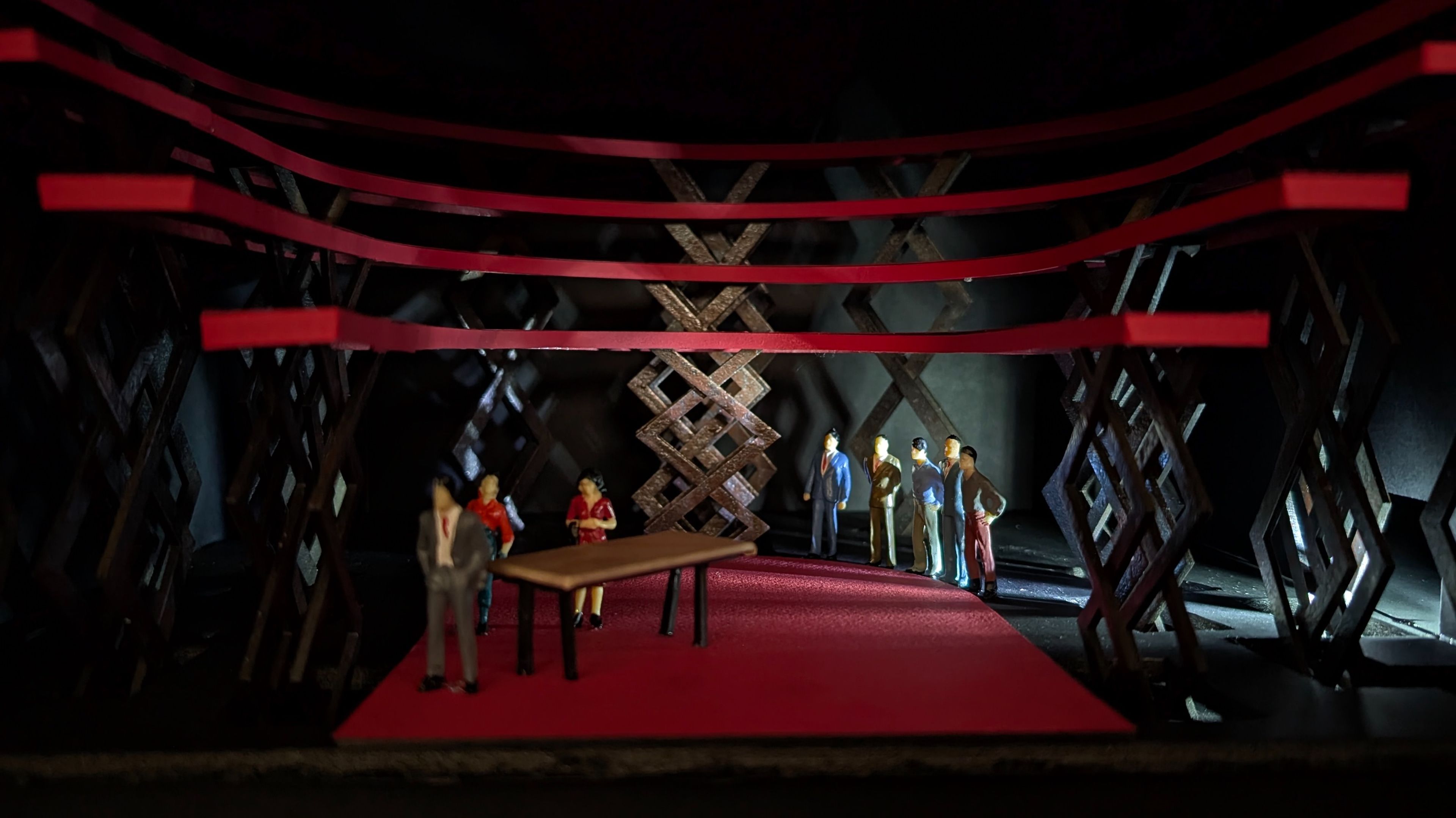 Final stage model showing the fully elevated set, with red platforms supported by scissor lift lattice structures and a group of miniature figures positioned around a table, highlighting the dynamic configuration.