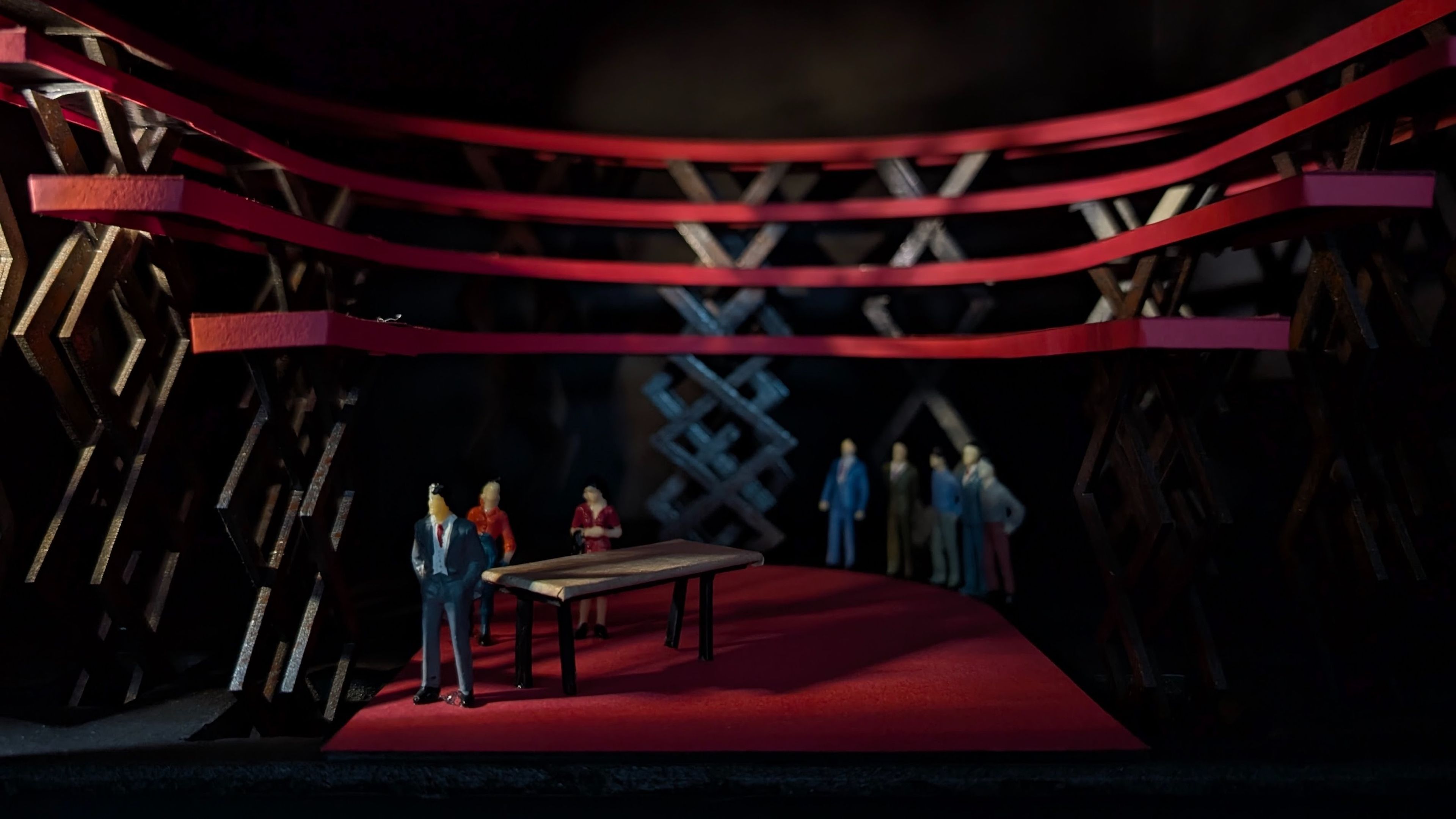 Scale model depicting the set design's transformation, with red platforms raised by scissor lift lattice structures. Miniature figures gather around a central table, emphasising the automated elevation of the set.