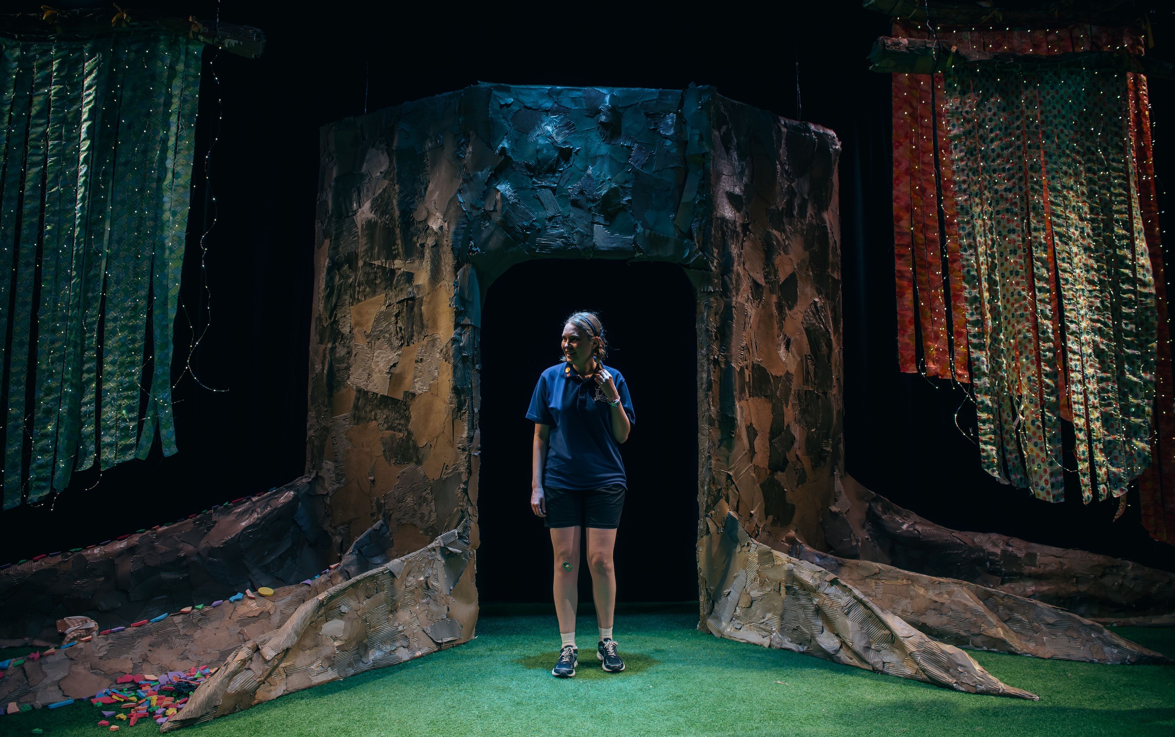 A performer stands in the centre of an abstract set featuring textured walls and colourful fabric panels, illuminated with green and orange lighting.