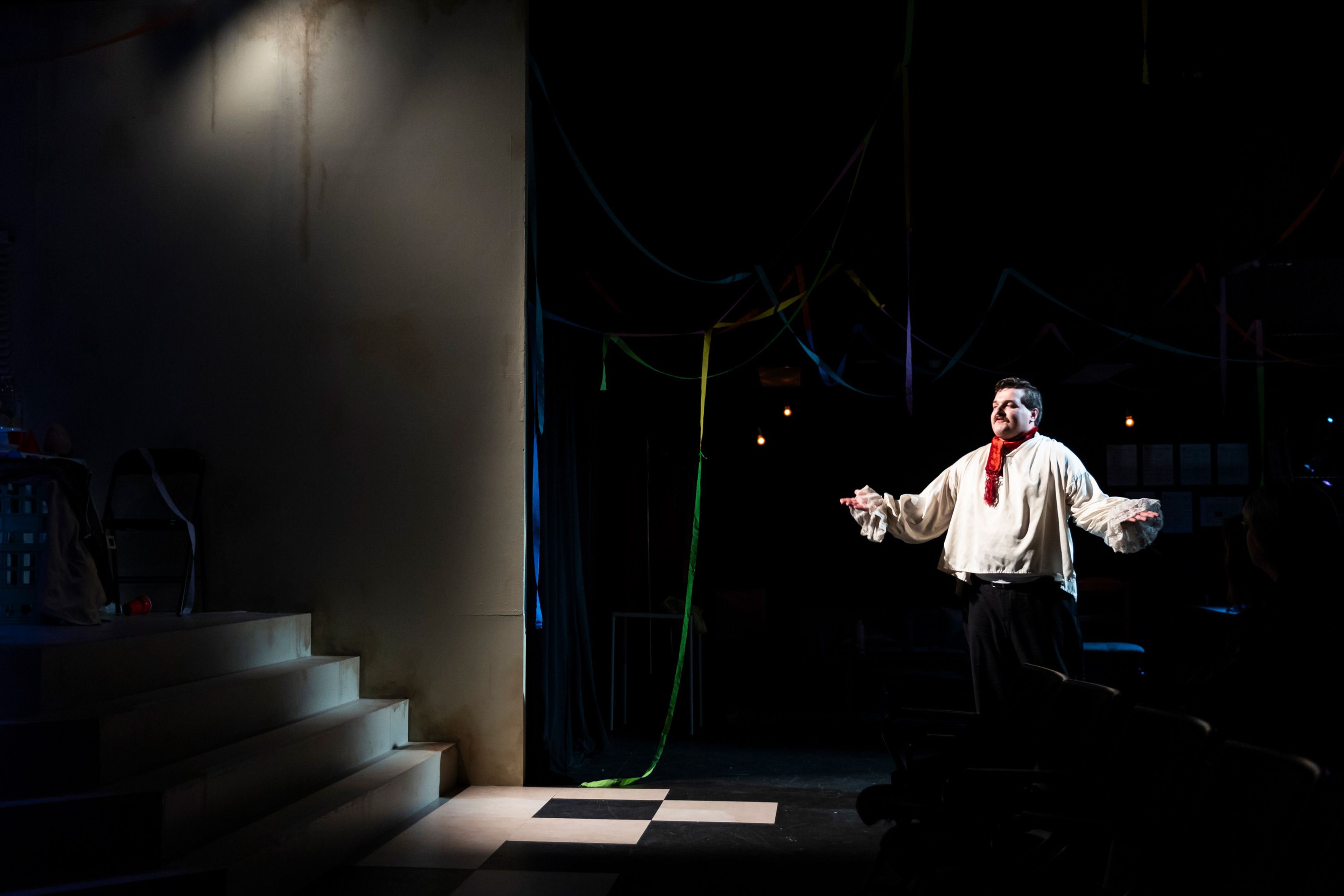 Theatre production photograph showing a performer in period-style white shirt with red cravat, dramatically lit against a checkerboard floor and colourful streamers.