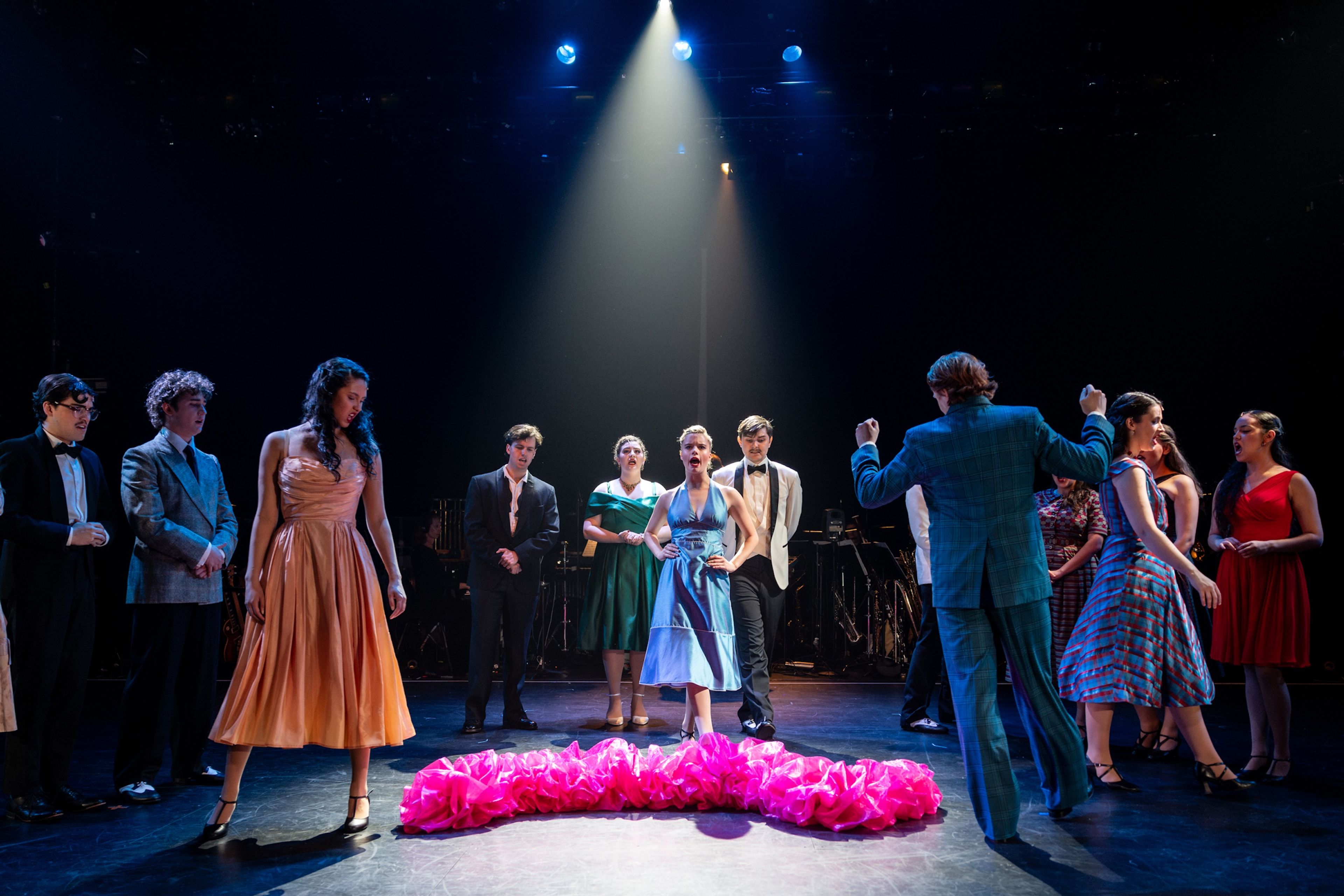 Theatre production photograph of a group of performers dressed in formal attire from various eras, standing in a spotlight with a bright pink ruffled costume piece lying on the stage floor.