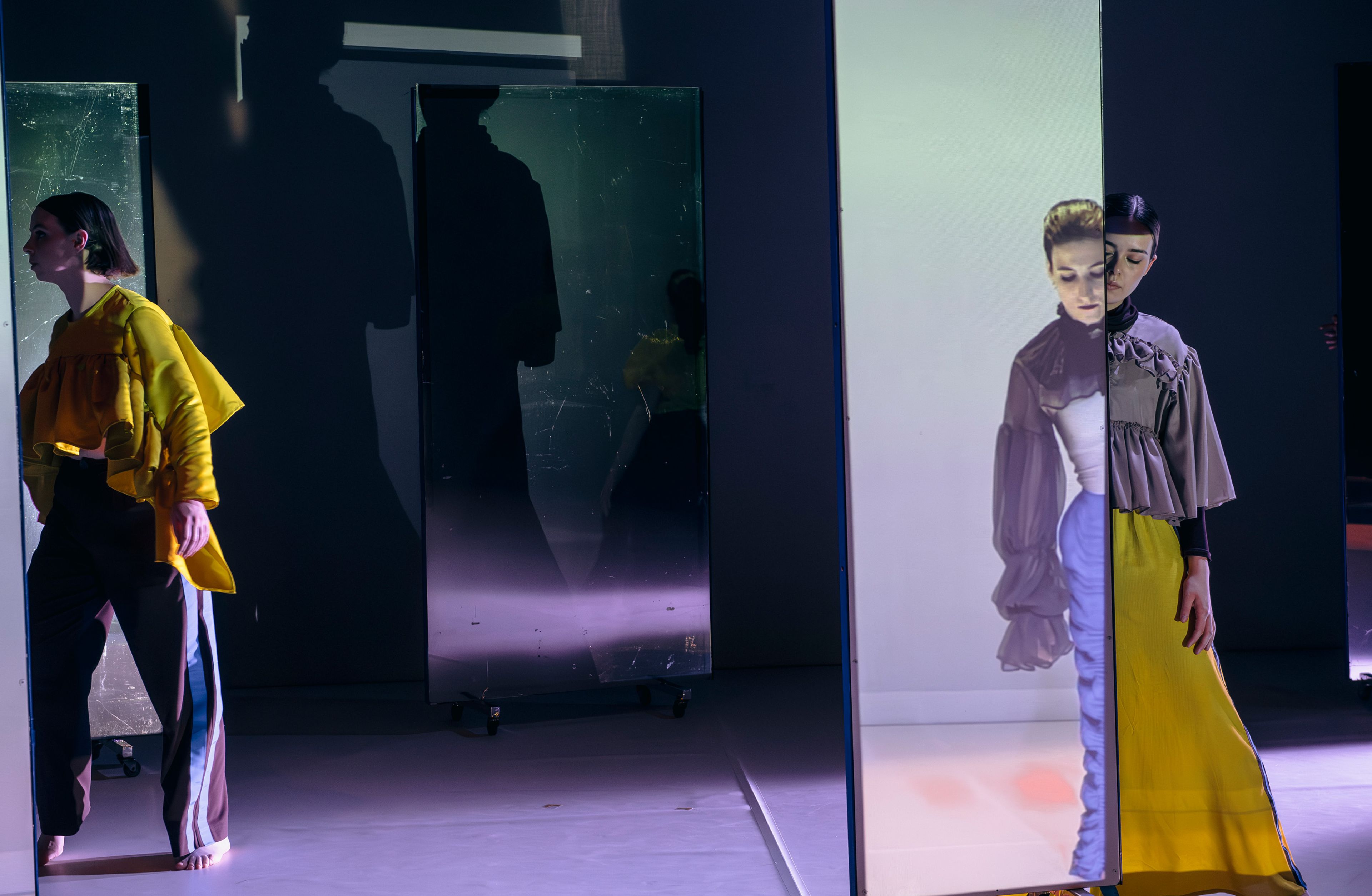 Dance production photograph featuring two dancers partially obscured by mirrors, casting shadows with bright, directional lighting creating a layered effect.