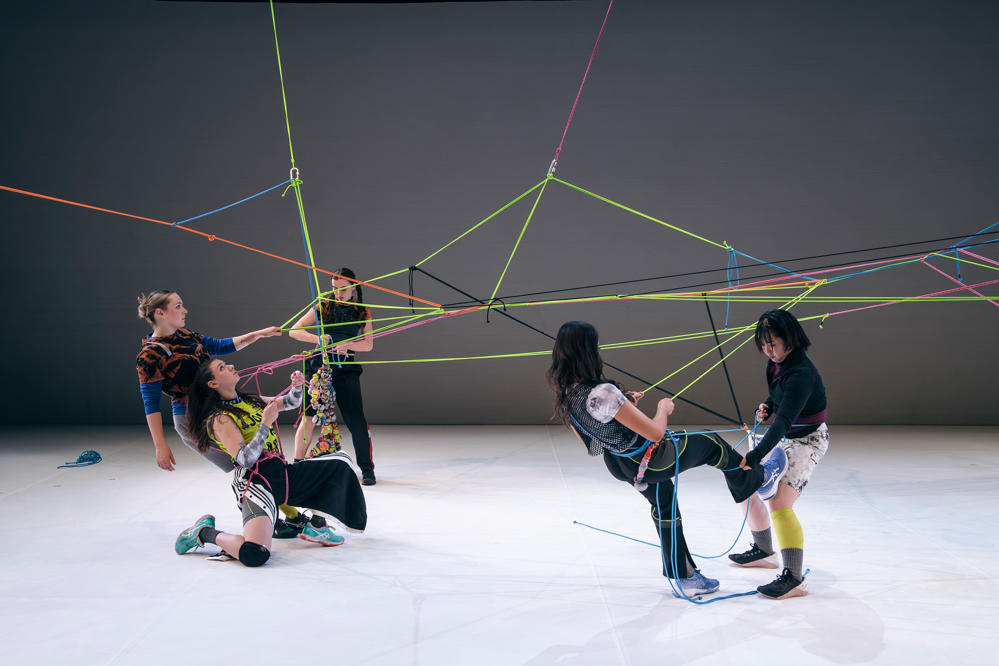 Dance production photograph featuring dancers interacting with neon-coloured ropes, creating a dynamic, geometric web on stage.