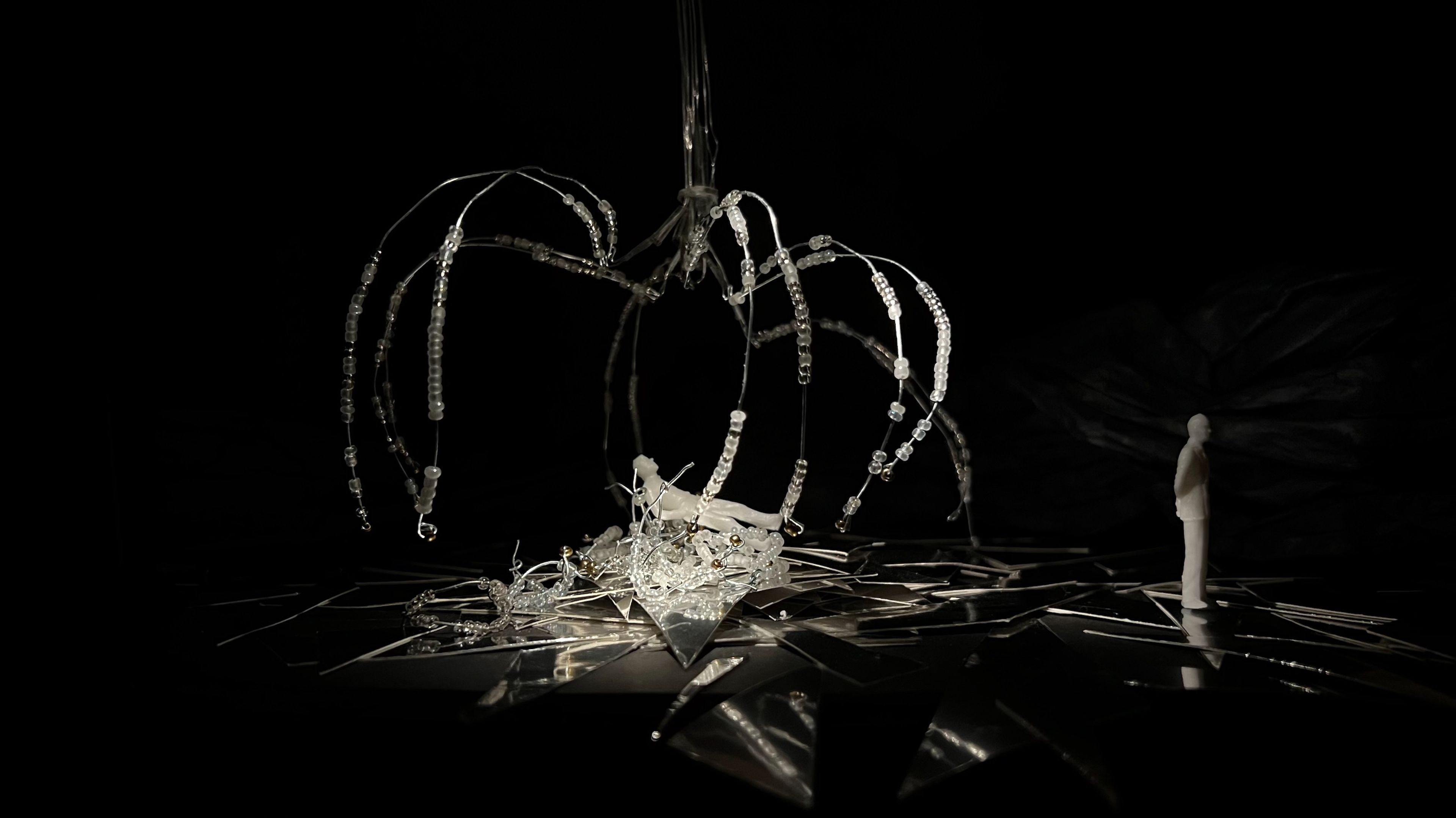 Theatre set model in monochrome, showing a white figure observing a sculptural arrangement of beaded strands and broken reflective surfaces against a black background