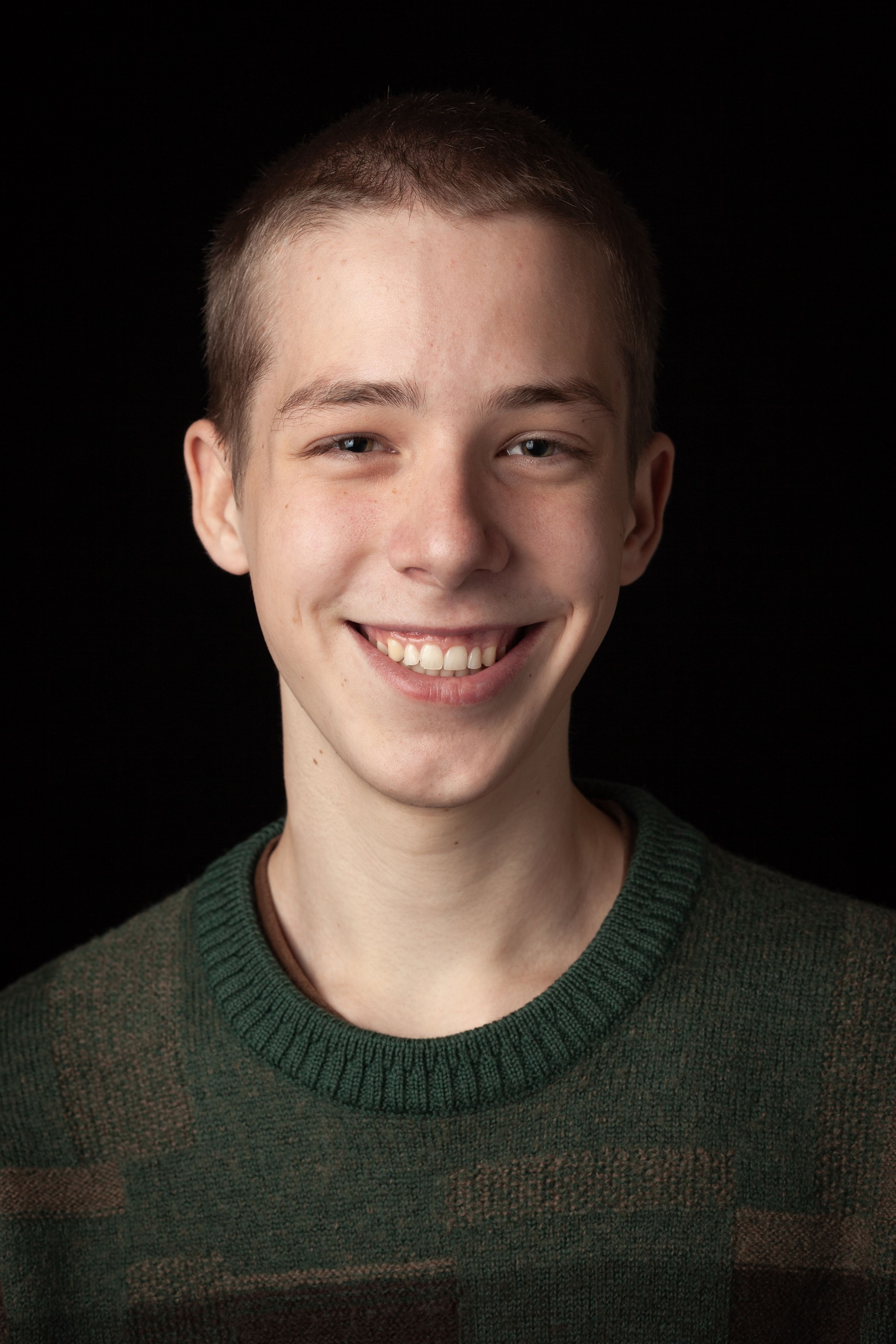 Headshot of a smiling young man with a close-cropped haircut, wearing a green patterned jumper, against a black background.