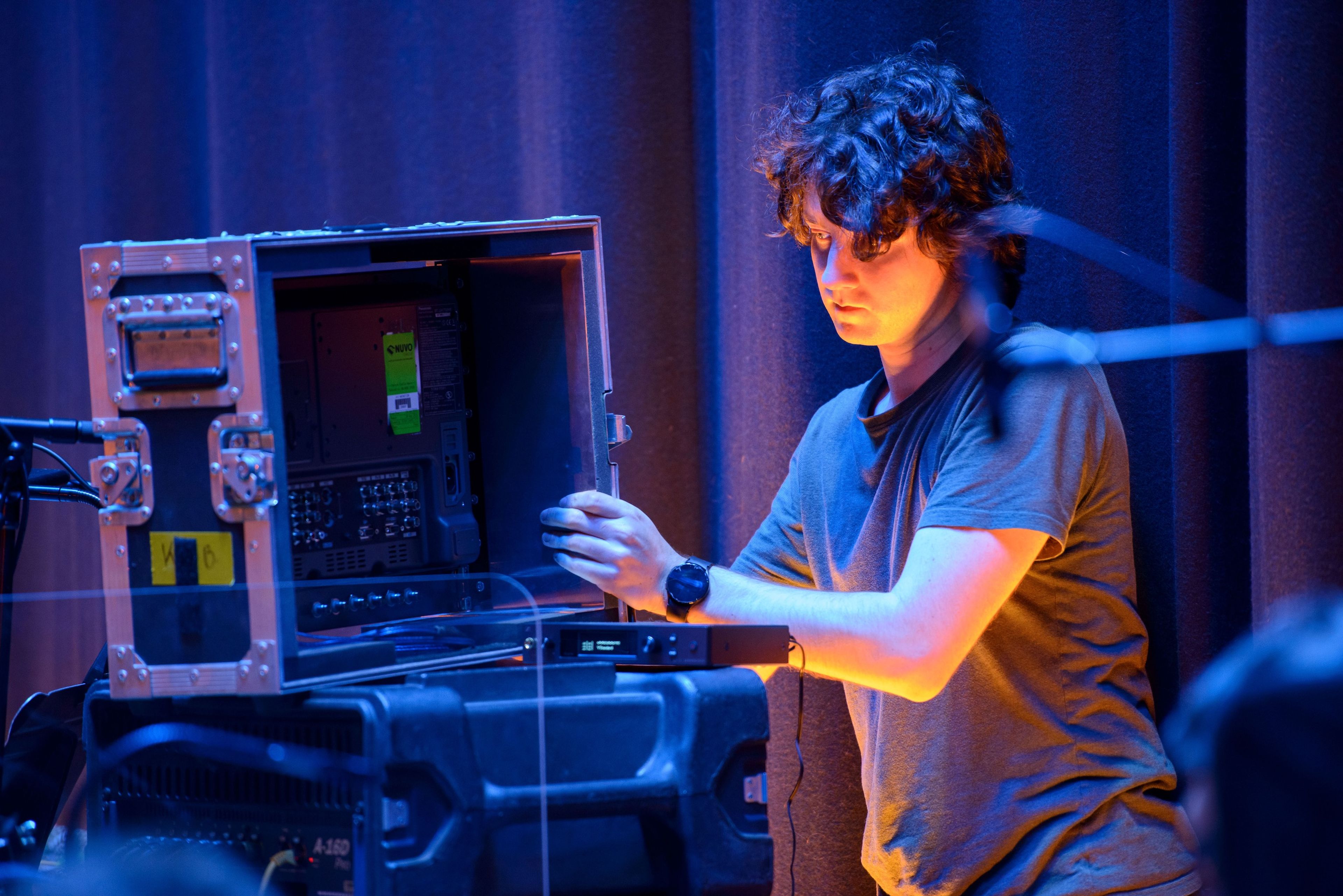 A person adjusts equipment inside a large metal case, with a focused expression, under blue stage lighting.