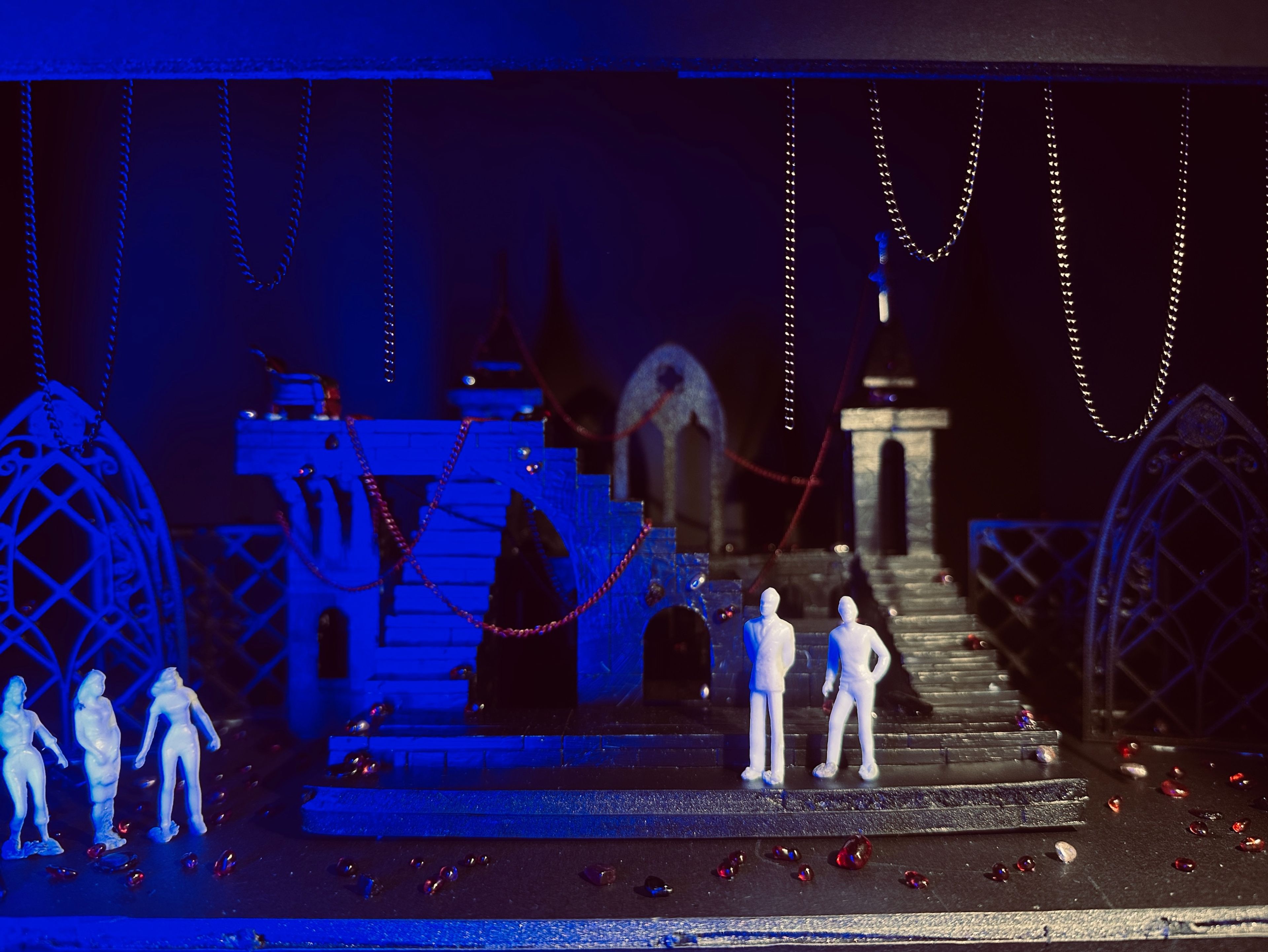 Gothic-style set model bathed in blue light, with white figures standing on staircases and platforms adorned with red beads and chains.