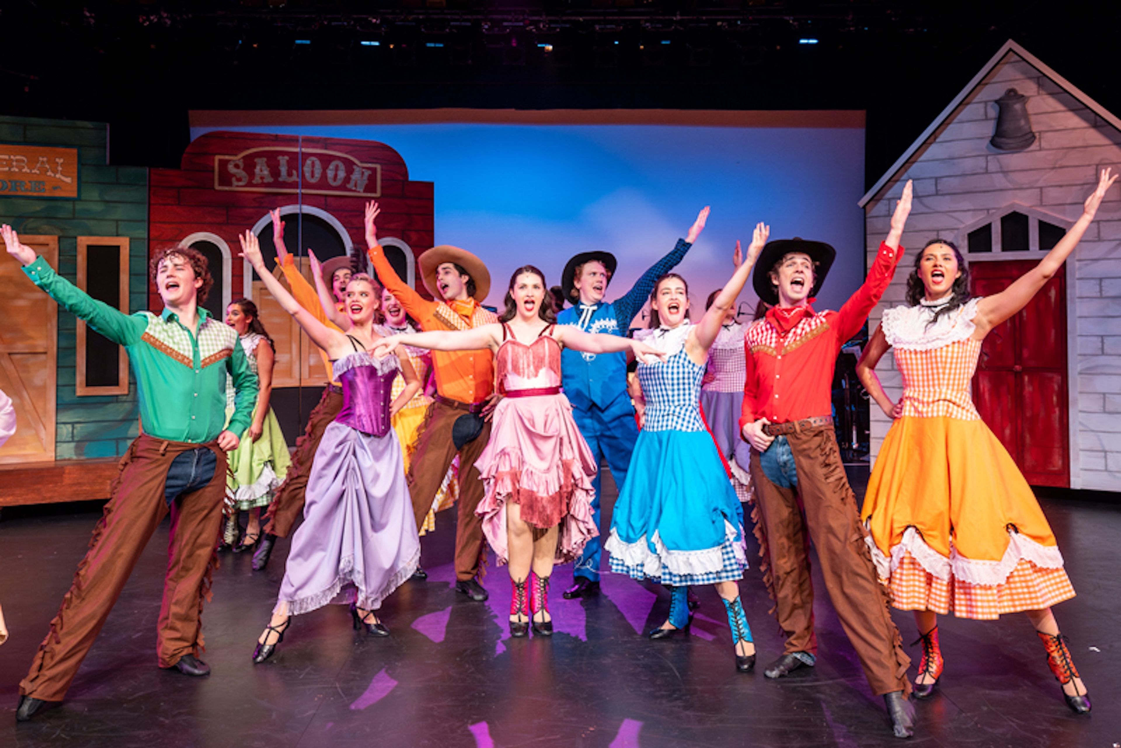 Production photograph of a Western musical number featuring performers in colourful period costumes including gingham dresses and cowboy attire, posed in front of a saloon set