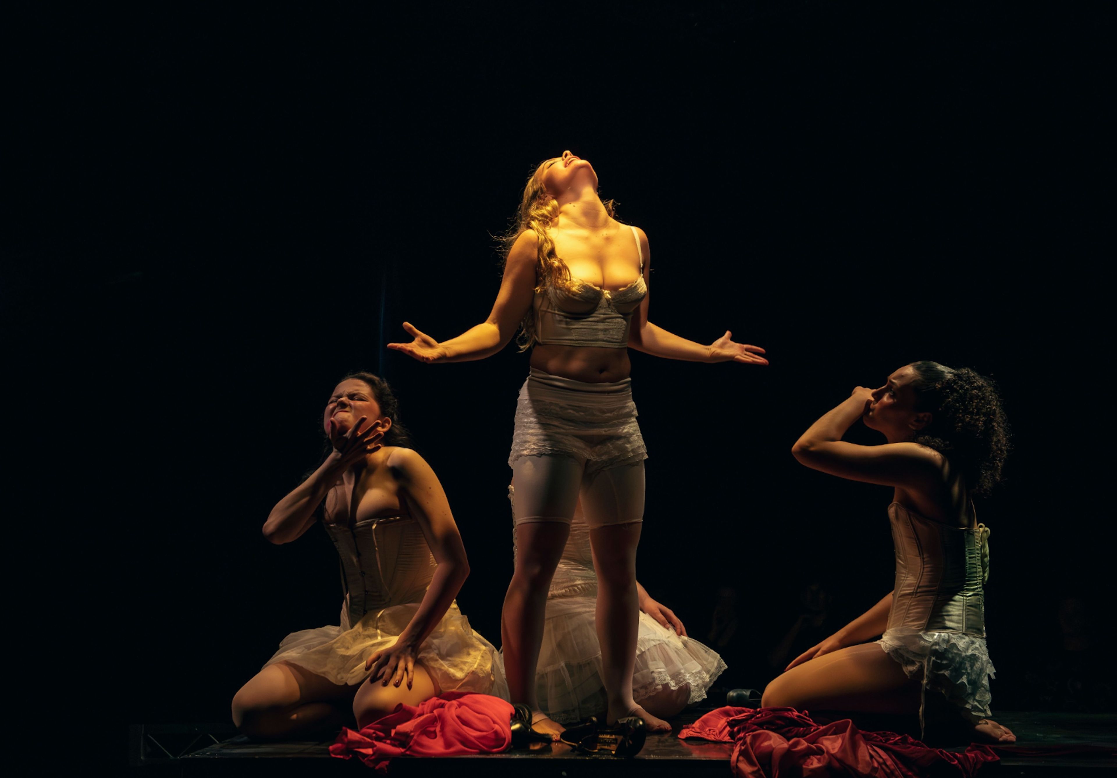 Theatre production photograph of three performers kneeling dramatically in white costumes with red fabric on the floor, illuminated by warm spotlighting against a dark background.