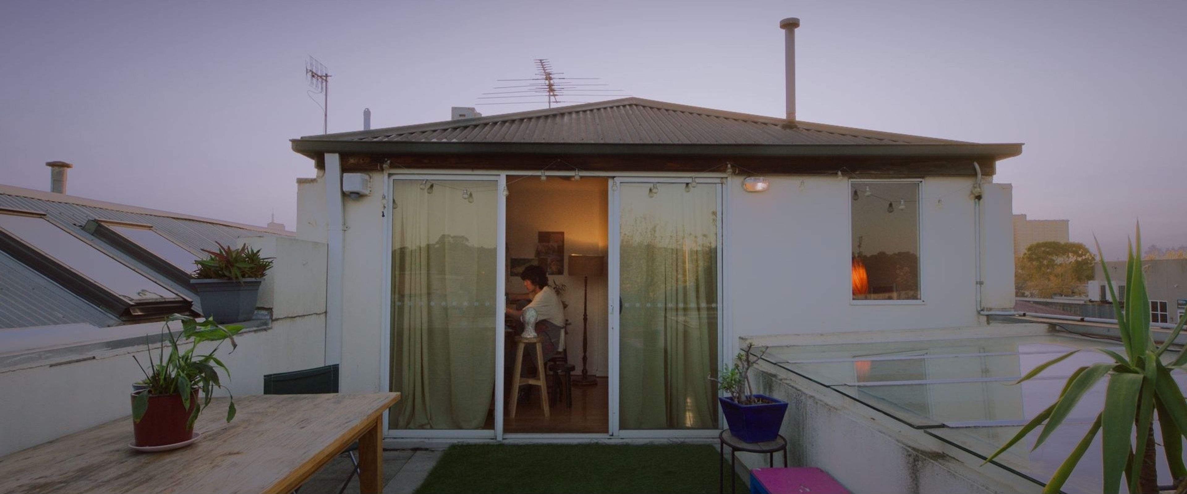 Film still of a rooftop apartment exterior at dusk, showing a view into an interior room where a person is seated and working.