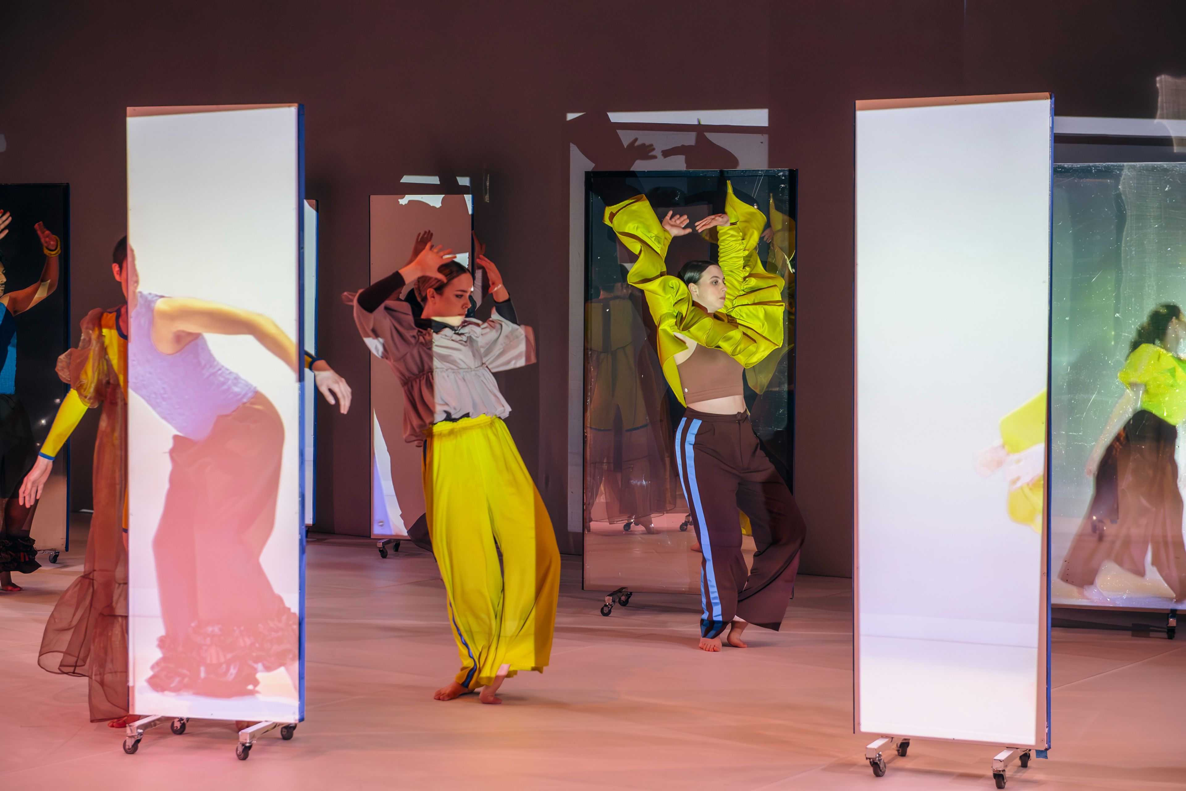 Dance production photograph of two dancers in dynamic poses, separated by movable mirrored panels, with colourful reflections cast on the stage