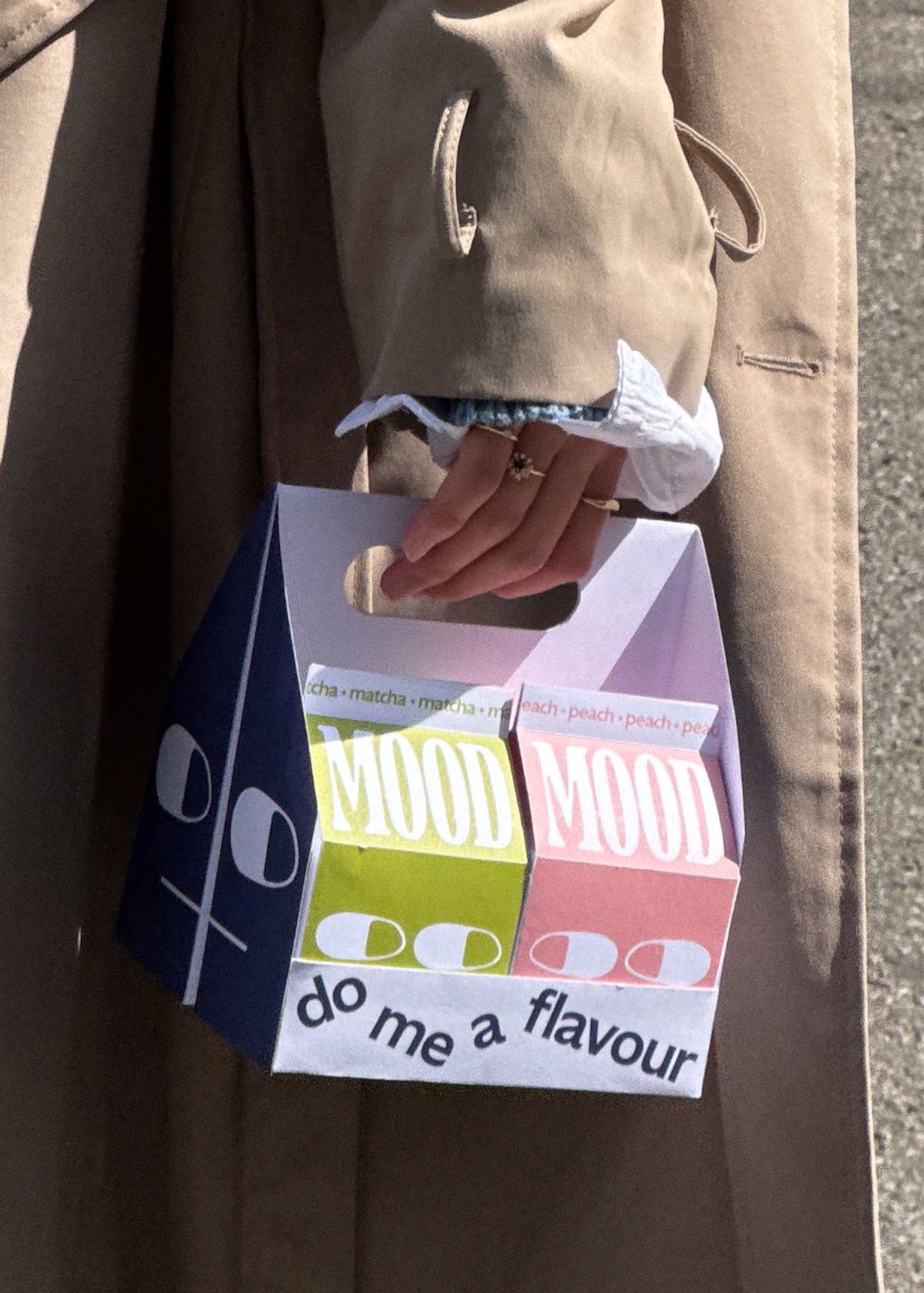 Paper carrier bag design for 'MOOD' beverages featuring multiple drink cartons in matcha green, peach pink, and navy colours. Text reads 'do me a flavour' beneath stylised faces made from pill capsule shapes. Photograph shows the bag being carried with a beige coat sleeve visible.