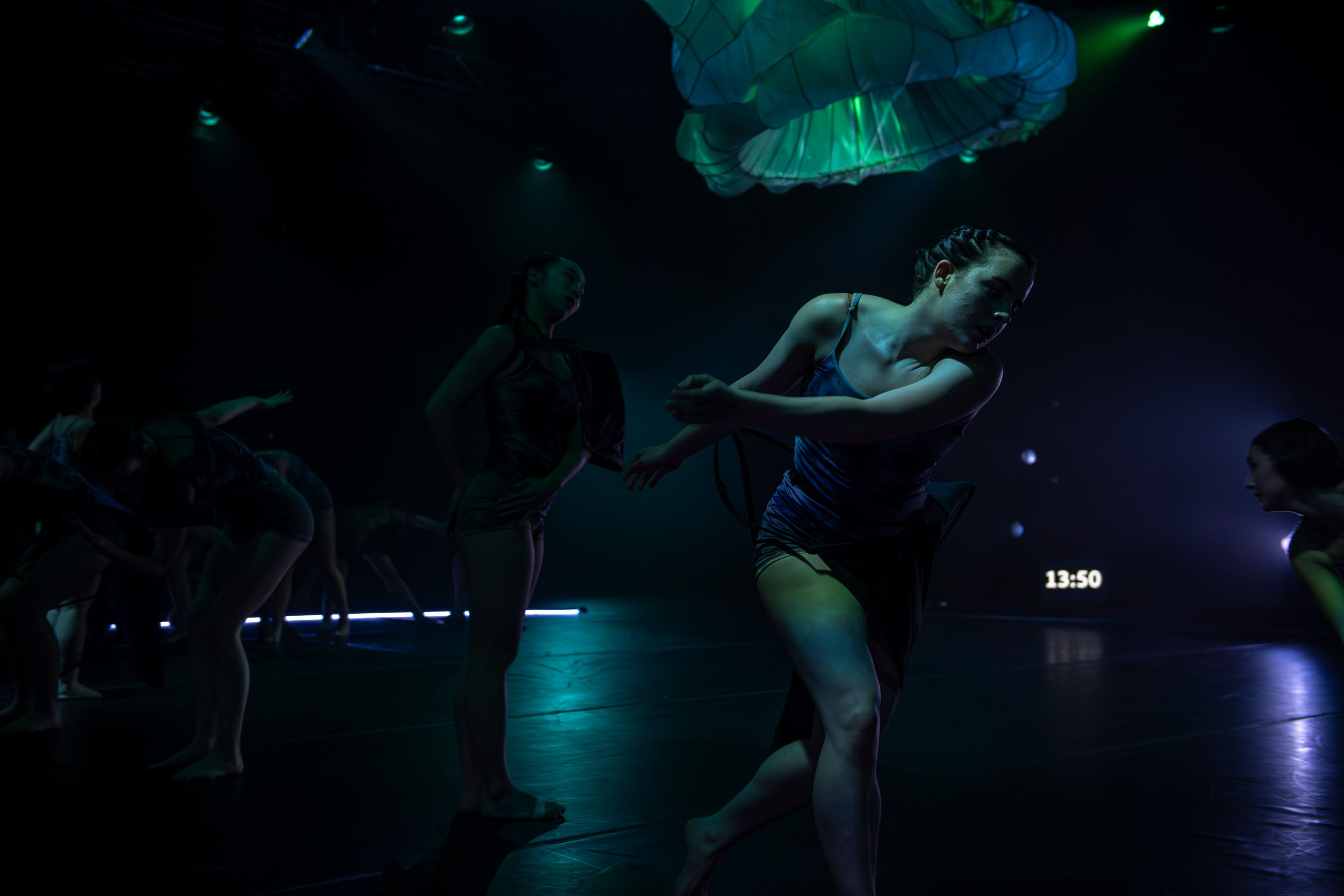 Theatre production photograph of dancers in dark costumes performing expressive movements, illuminated by green light with white fabric sculptures overhead.