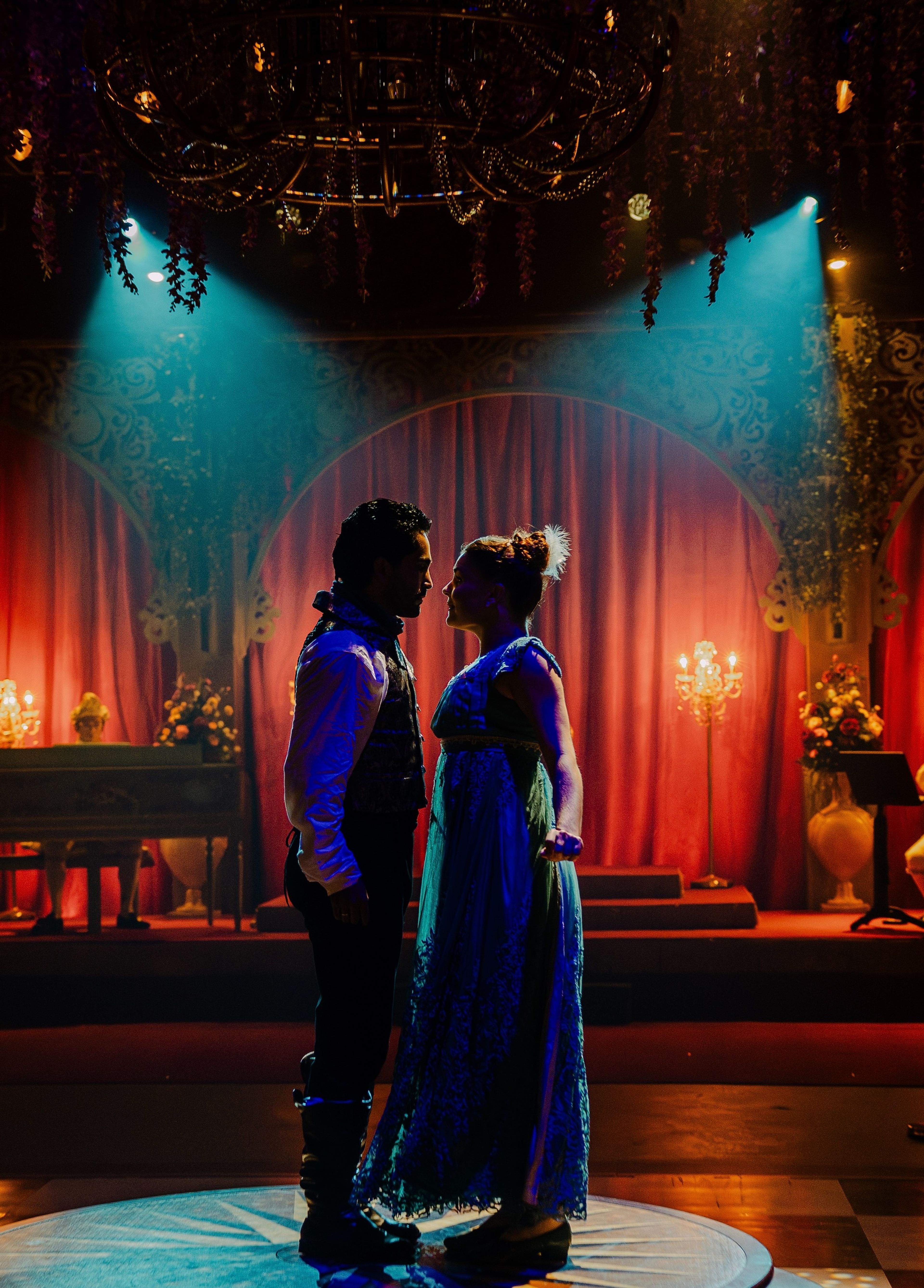 Theatre production photograph of two characters in period costumes standing close together in a romantic, warmly lit, ornate set with chandeliers and rich drapery.