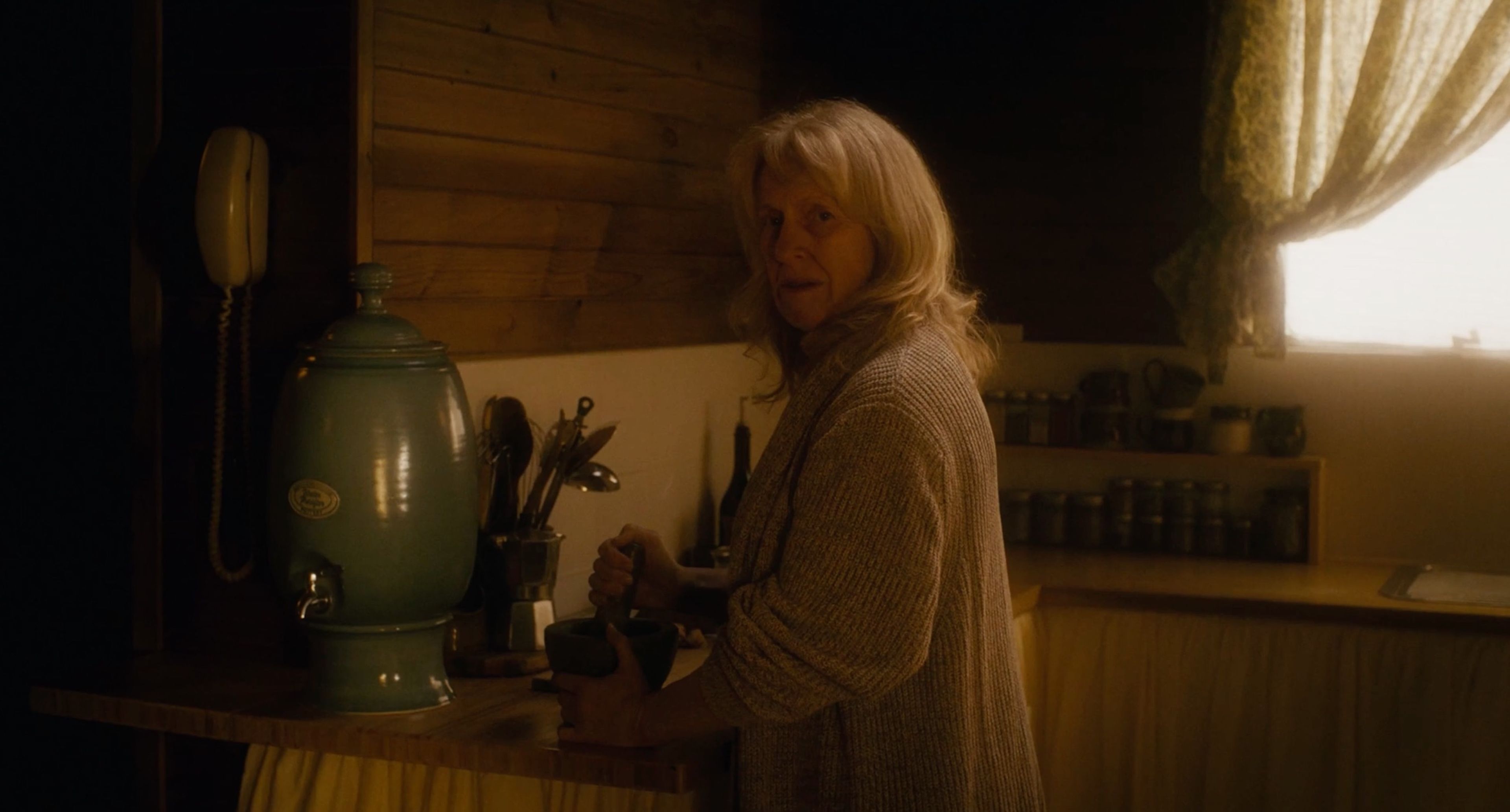 Film still depicting a woman in a beige cardigan in a rustic kitchen, lit warmly, with a large mint-green water dispenser and kitchen implements visible on wooden benchtops.