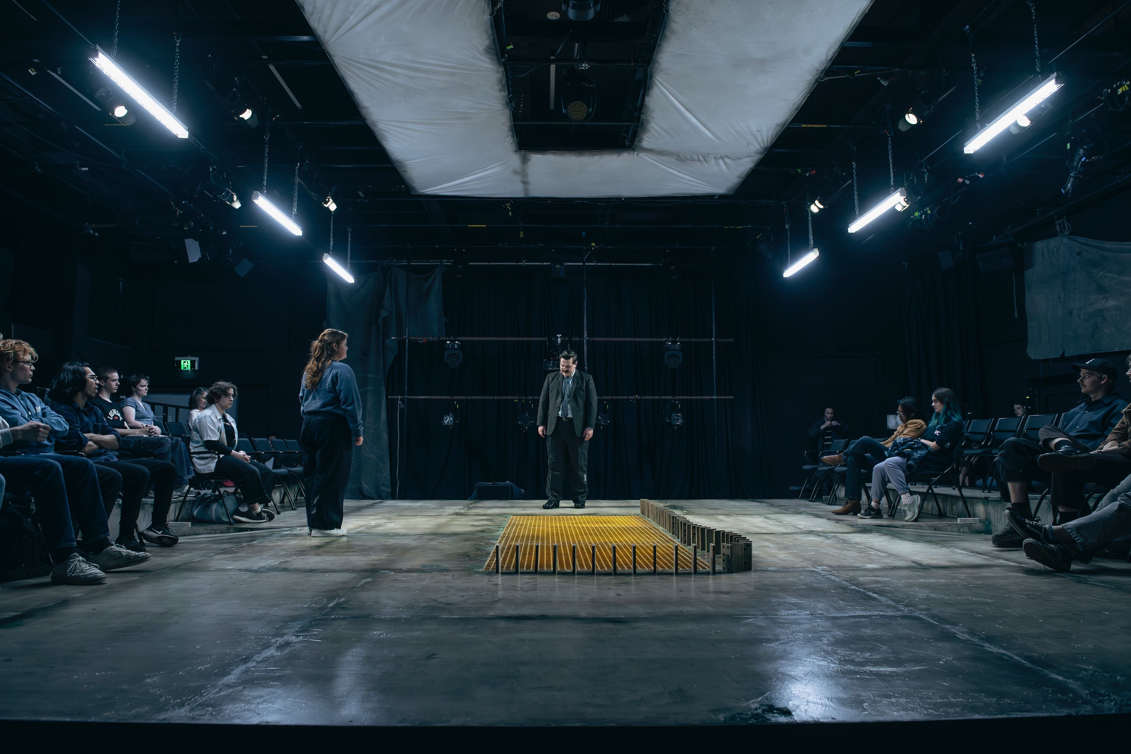 Theatre photograph showing a thrust stage with performers and audience arranged in traverse, featuring fluorescent lighting grid and a yellow central rectangle