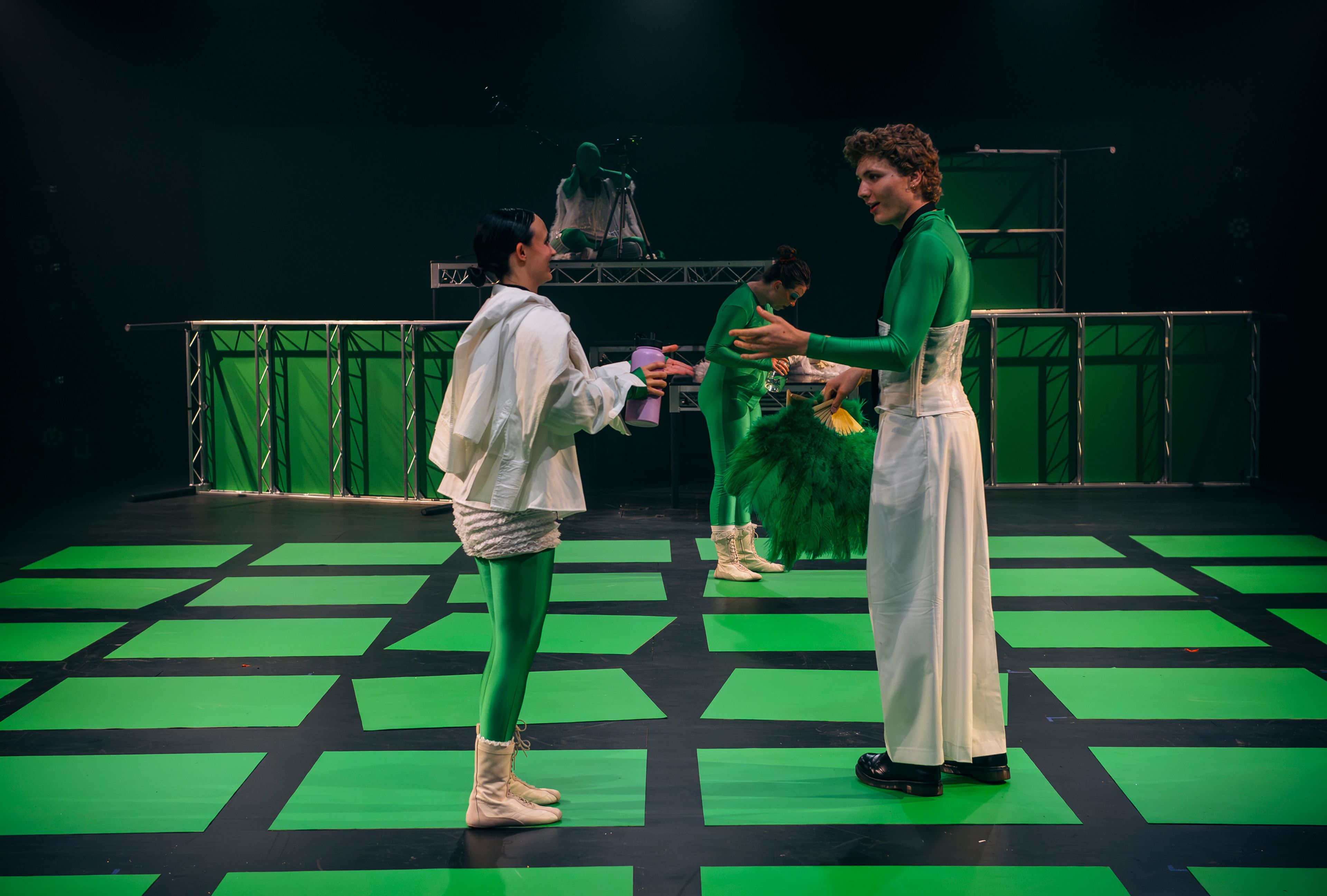 Theatre production photograph featuring actors in green costumes conversing on a stage with a bold green and black grid pattern, with green panels and metallic frames as part of the set.