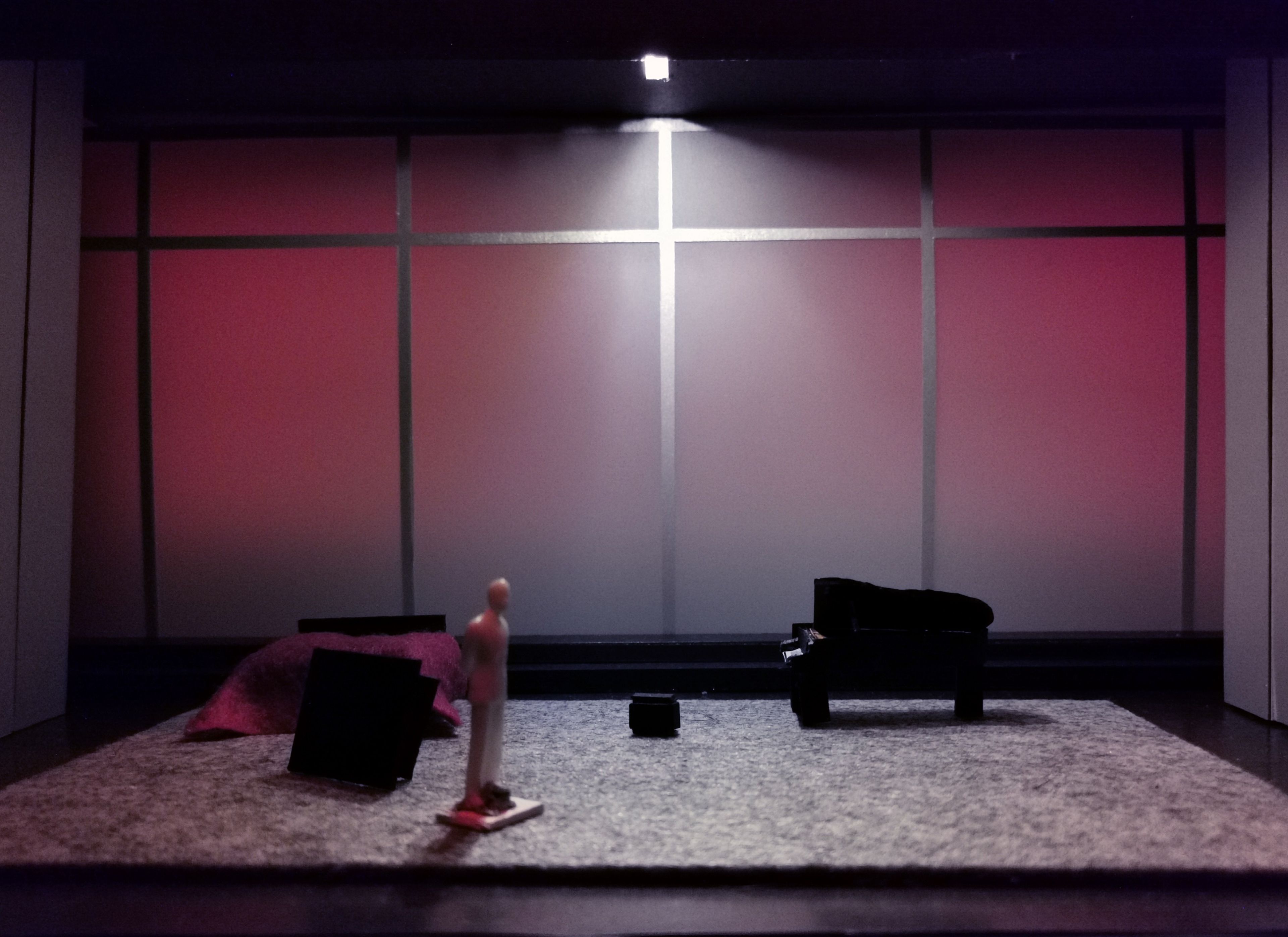 Scale model of a set design featuring a figure against a backdrop with a soft pink glow, with dark furniture scattered around.