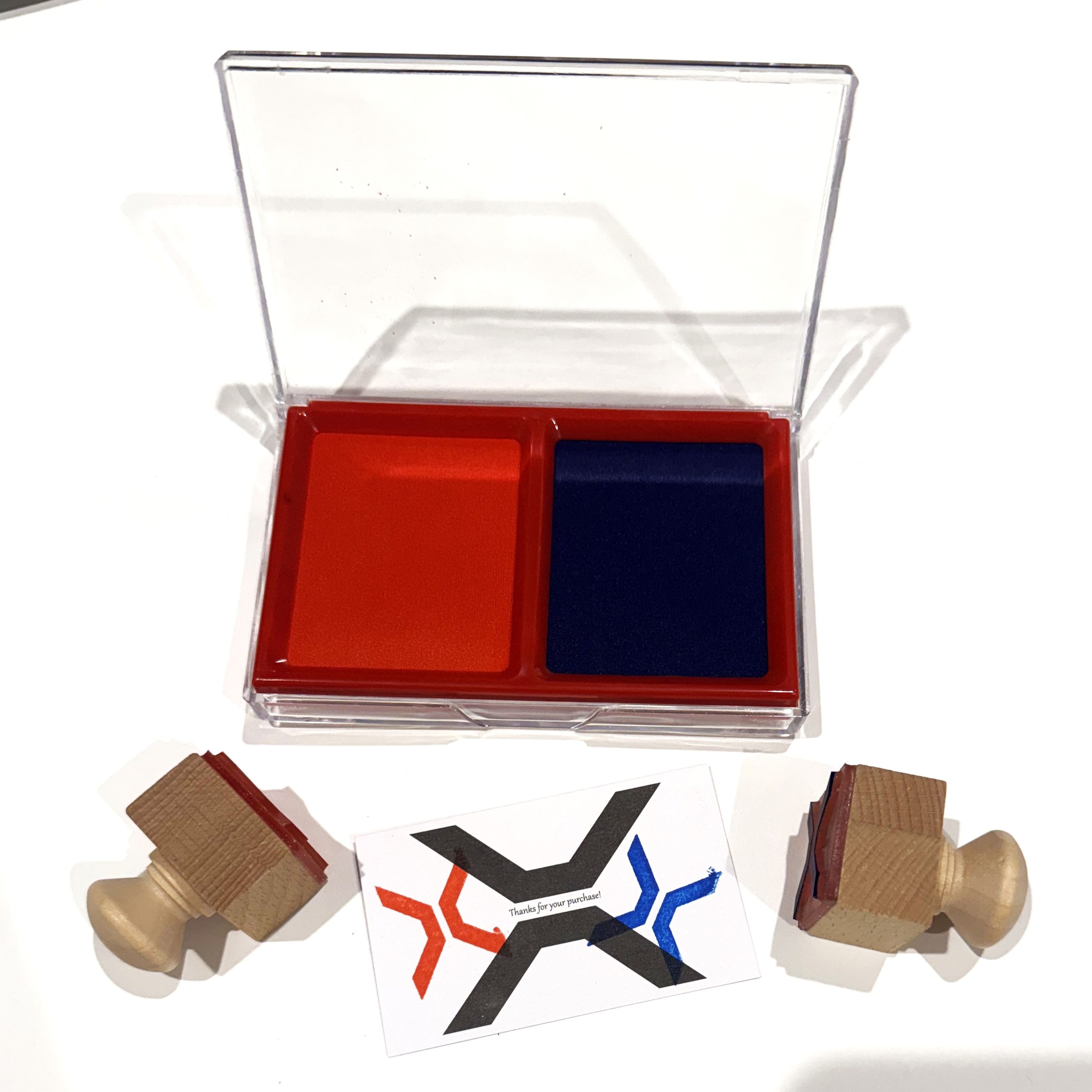 Two wooden stamps and a red and blue ink pad set, accompanied by a printed card featuring a stylised "X" symbol. The objects are arranged on a white background, emphasising the branding colours of red and blue.