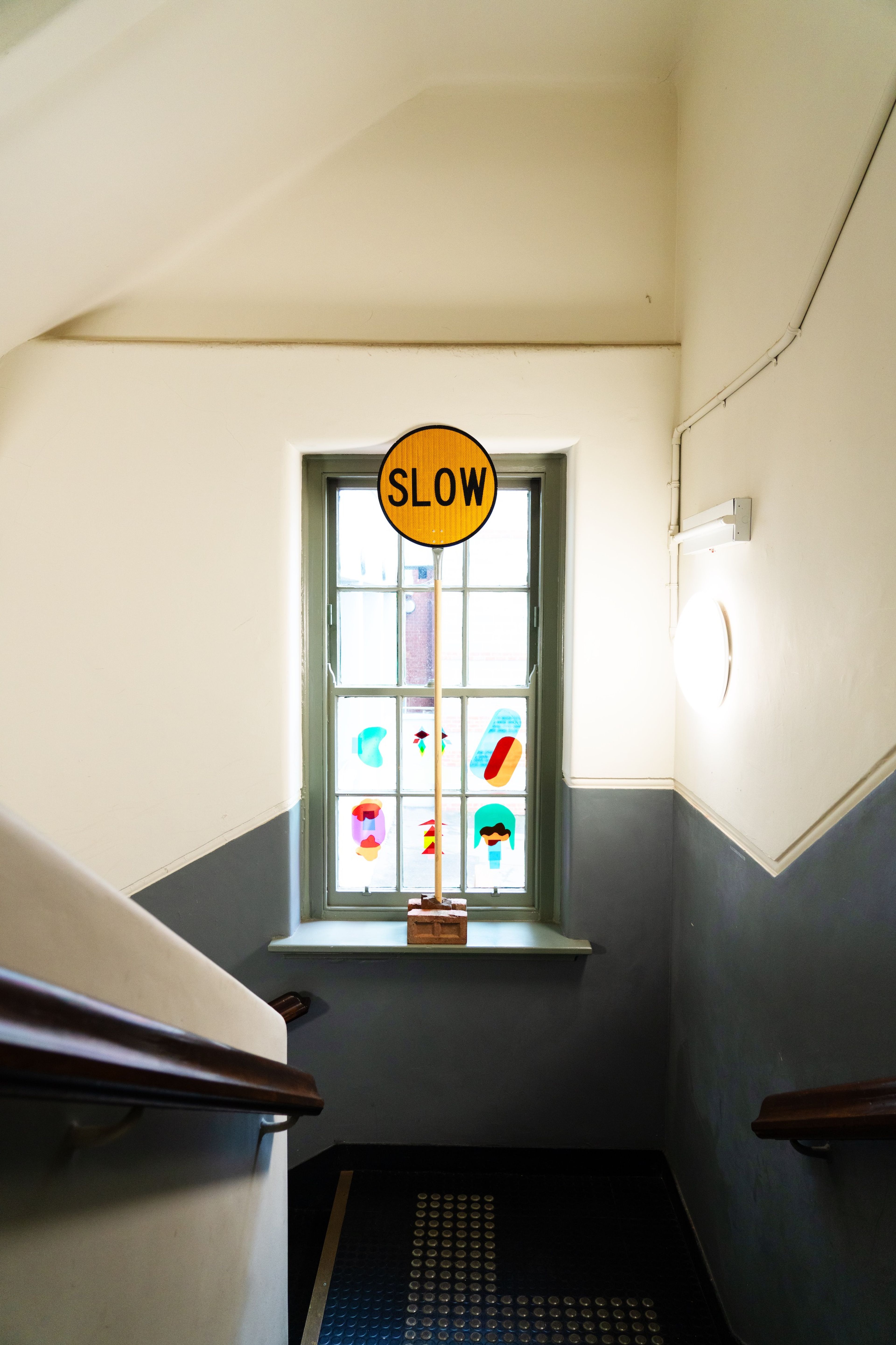 A stairwell features a "SLOW" traffic sign placed against a window with colorful abstract decals. The setup creates a blend of urban and playful aesthetics in an unexpected space.