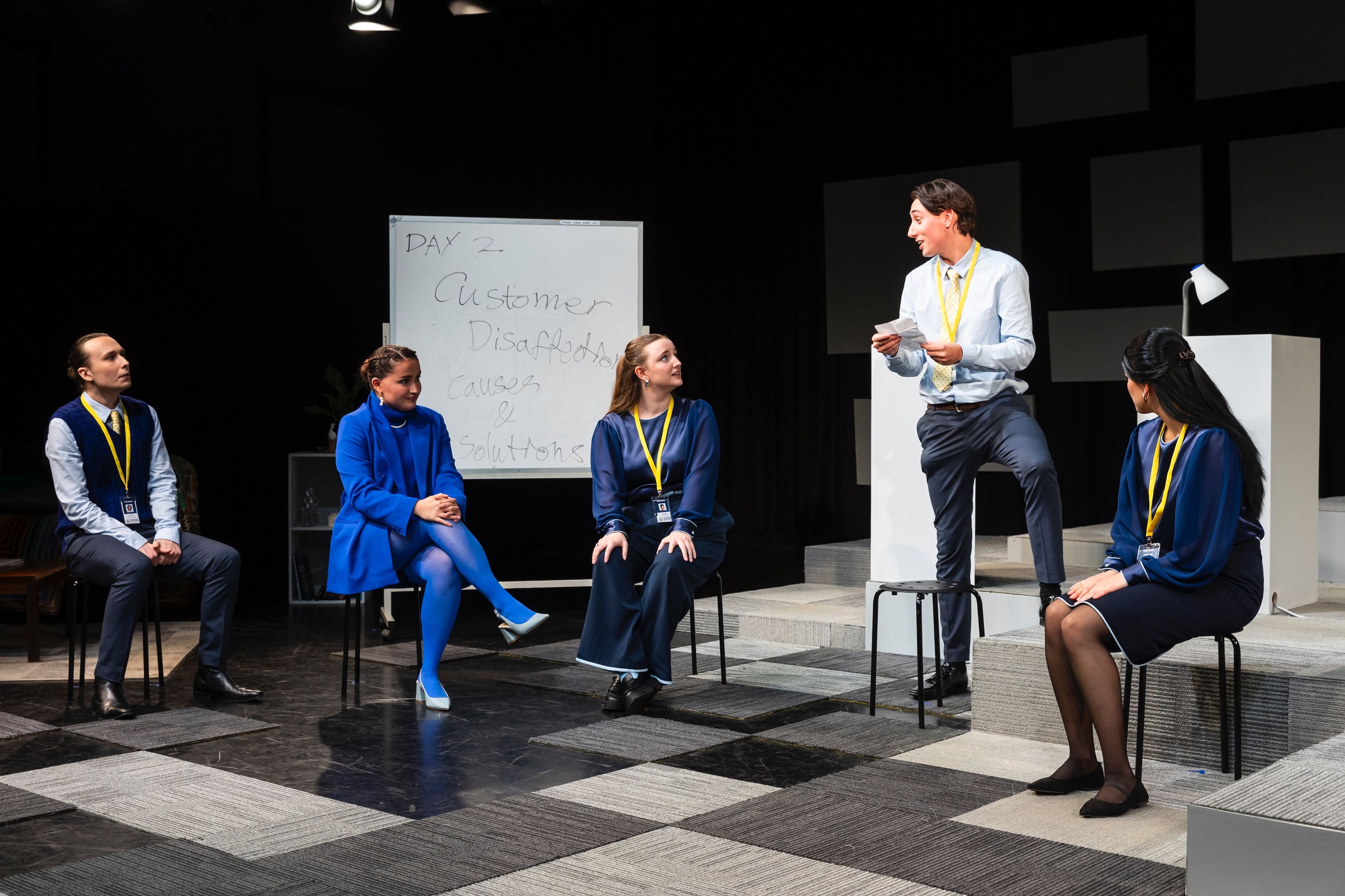 Theatre production photograph of actors in a corporate training setting, with a whiteboard reading “Day 2: Customer Disaffection – Causes & Solutions”.