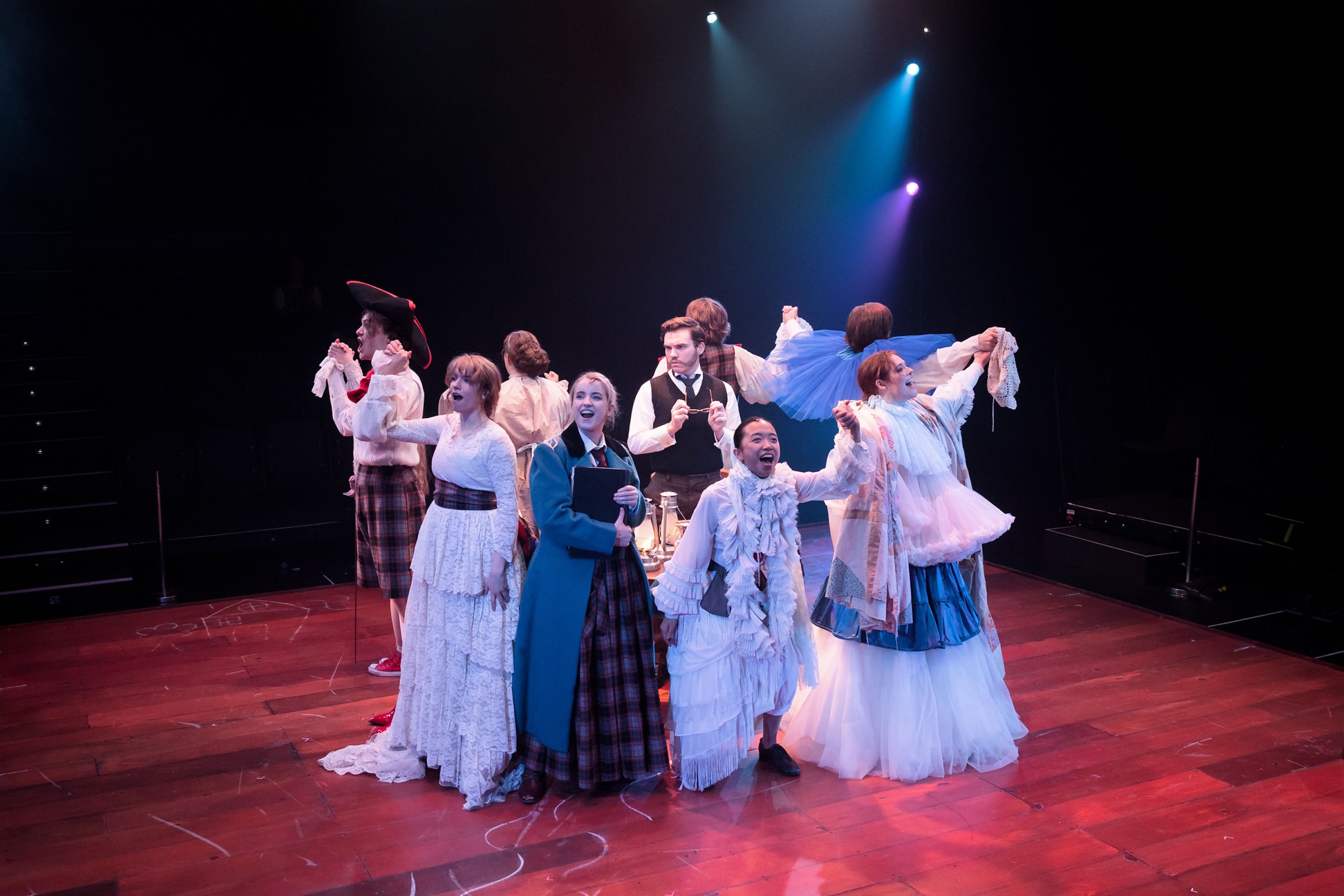 Theatre production photograph of a group of performers singing or celebrating, dressed in a mix of period costumes, with purple and blue stage lights adding atmosphere to the scene.