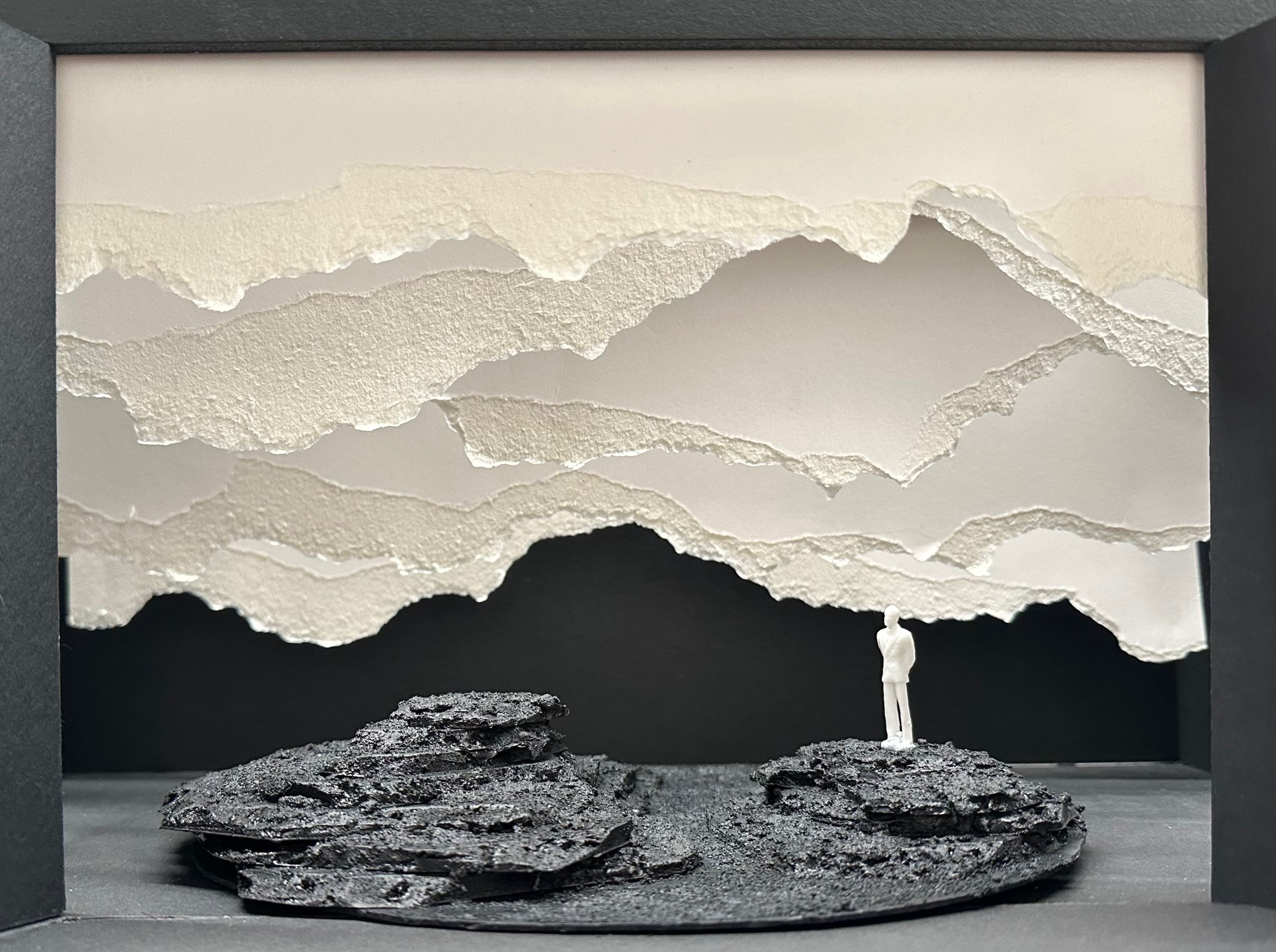 Scale model set design featuring layered paper mountains and textured rock formations in the foreground, viewed in neutral lighting.