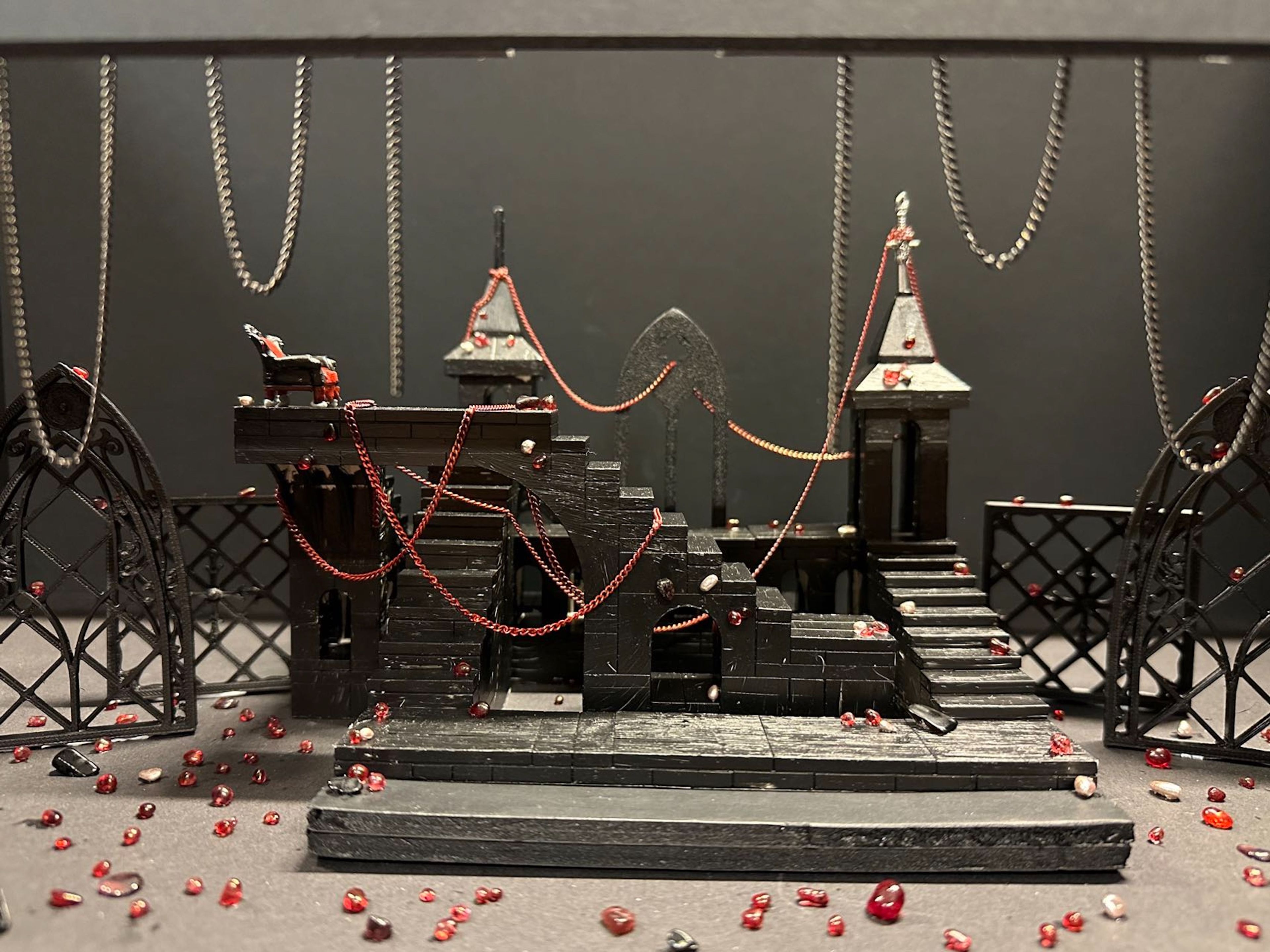 Scale model of a gothic-inspired set design with chains hanging above and red beads scattered around, featuring gothic arches and staircases.