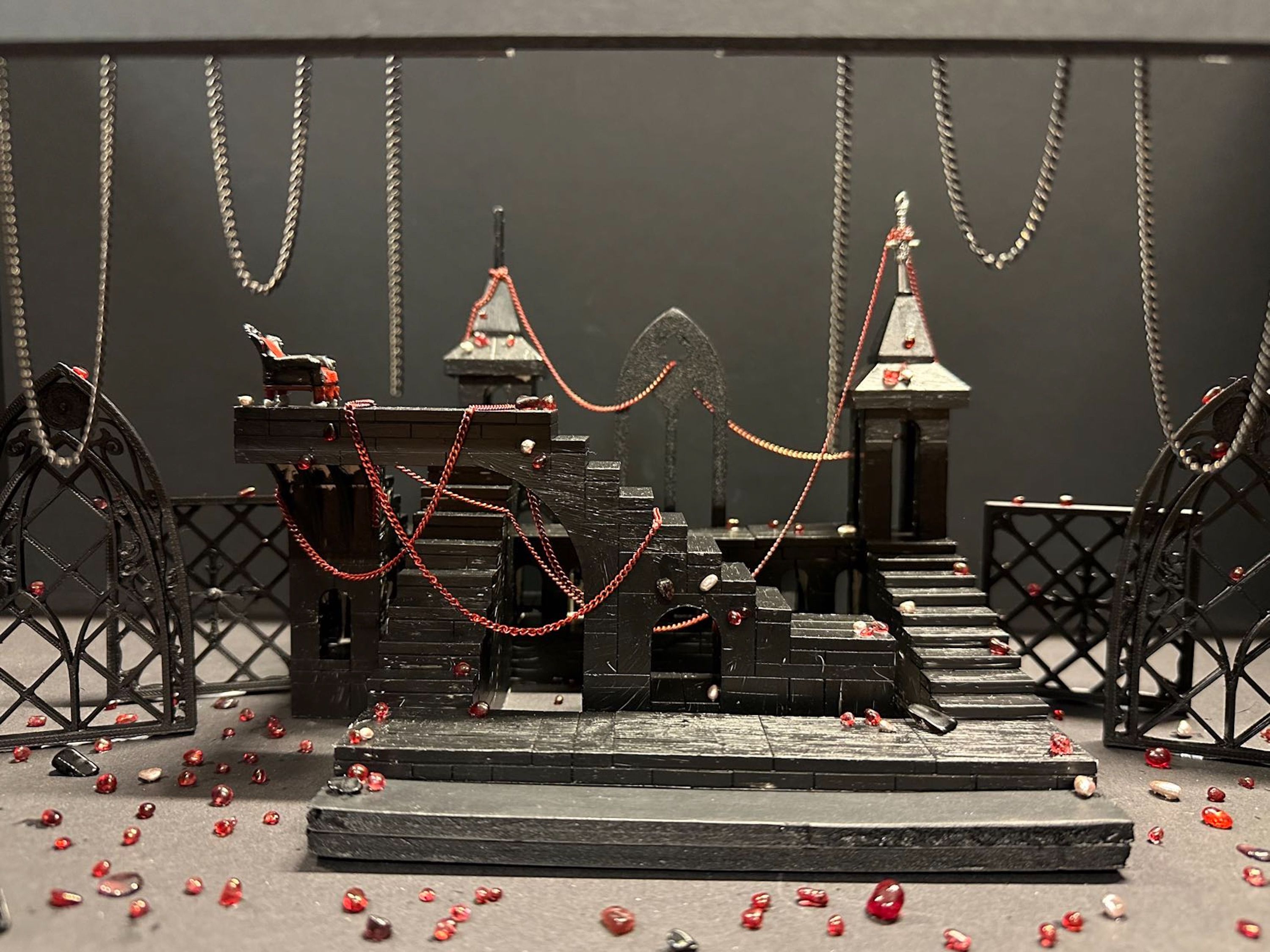 Scale model of a gothic-inspired set design with chains hanging above and red beads scattered around, featuring gothic arches and staircases.