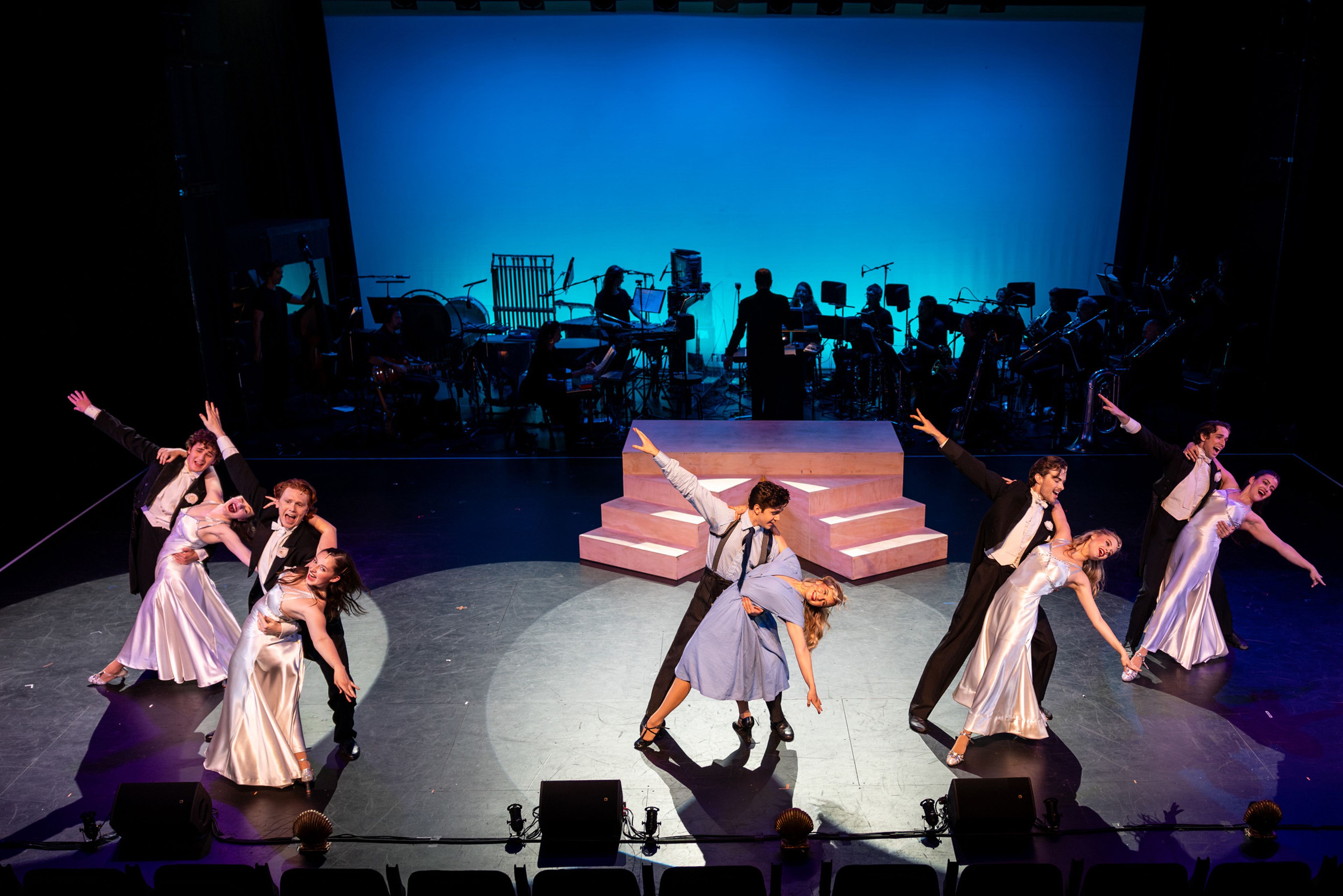 Theatre production photograph showing couples performing a formal ballroom dance number, with women in satin gowns and men in black tie, against a silhouetted orchestra and stepped set piece.