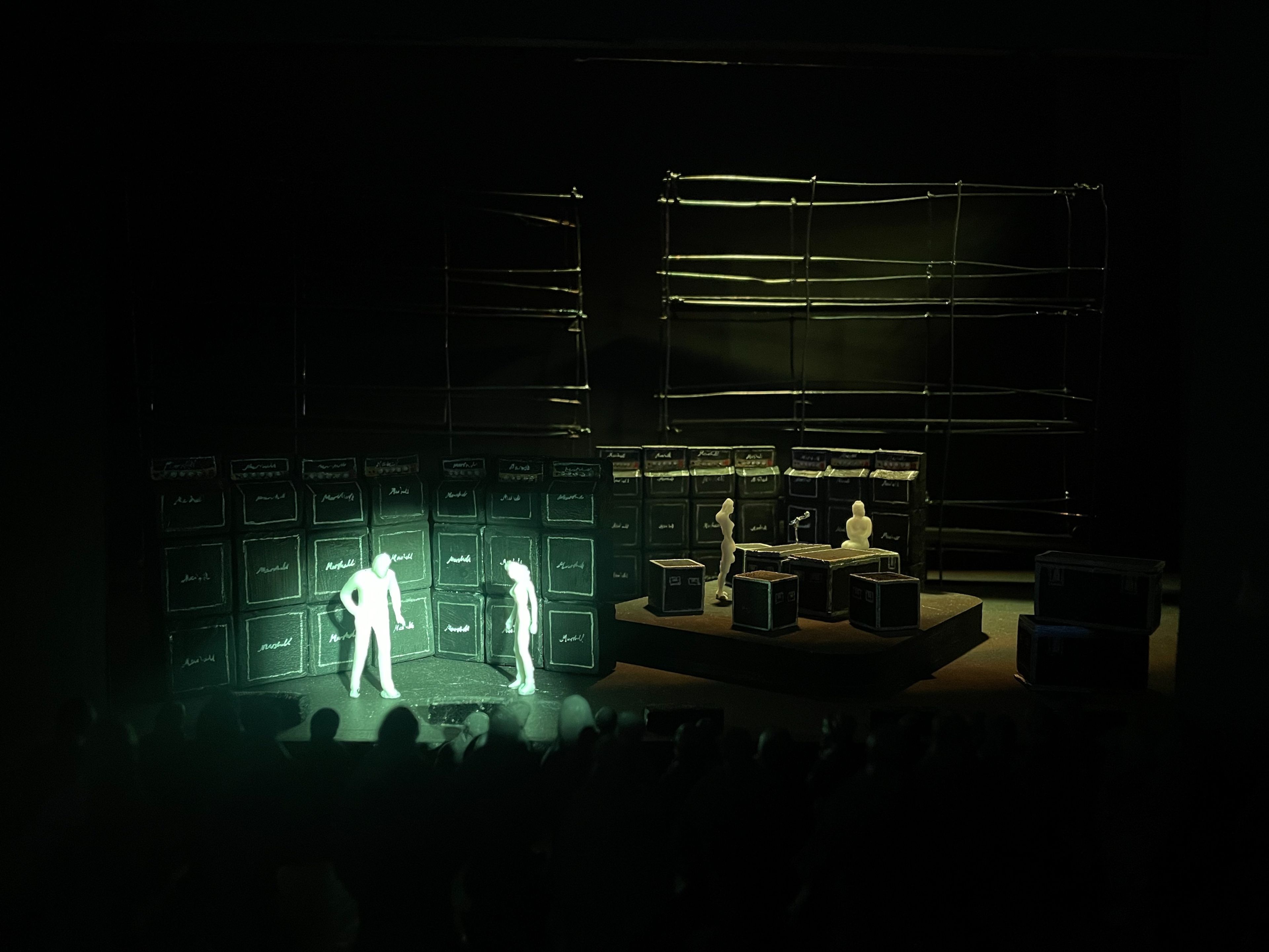 Scale model of a set design, showcasing two figures in front of a wall of amplifiers illuminated in green.