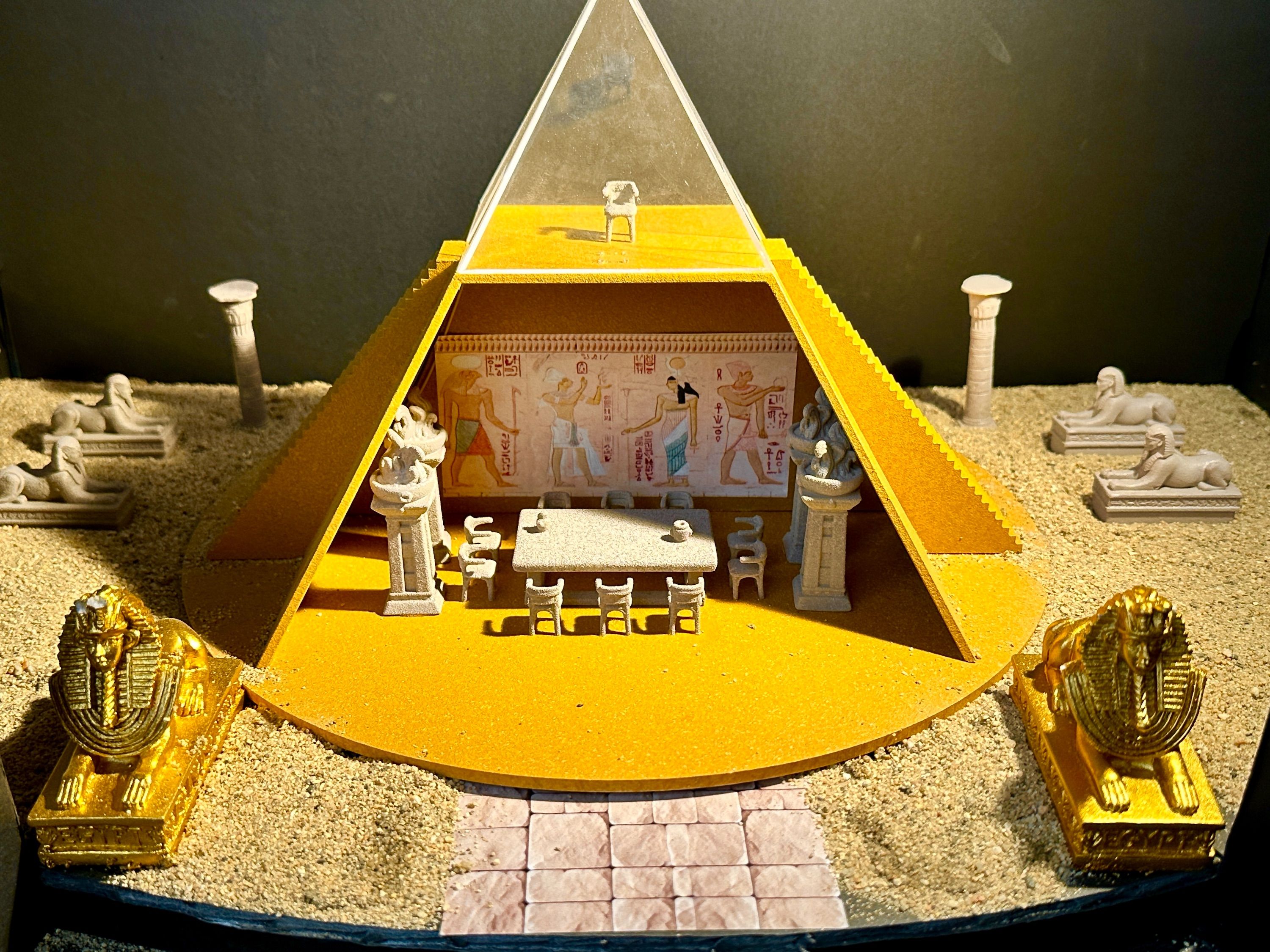 Scale model of an Egyptian tomb interior with gold pyramid entrance, hieroglyphic wall art, miniature furniture, and sphinx statues flanking a tiled pathway