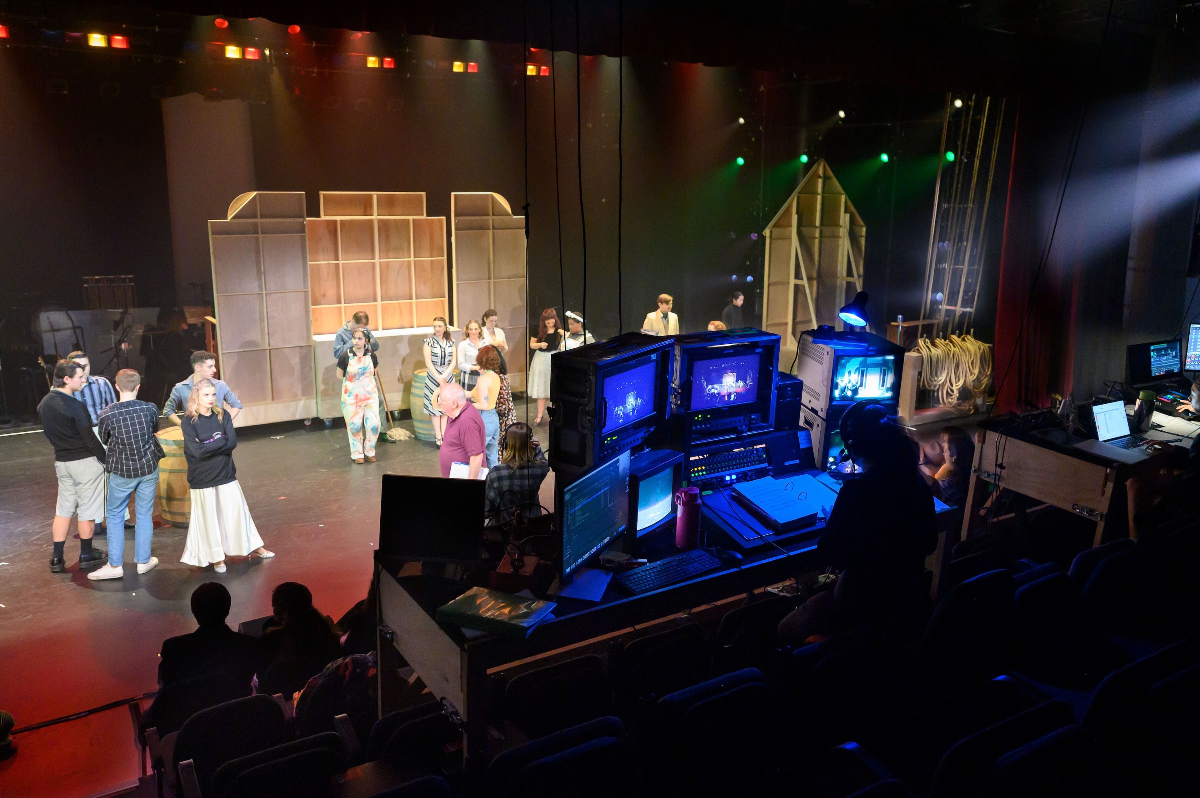 View from the technical booth looking towards the stage, where actors gather and rehearse in costume, with control screens and equipment in the foreground showcasing the behind-the-scenes setup.