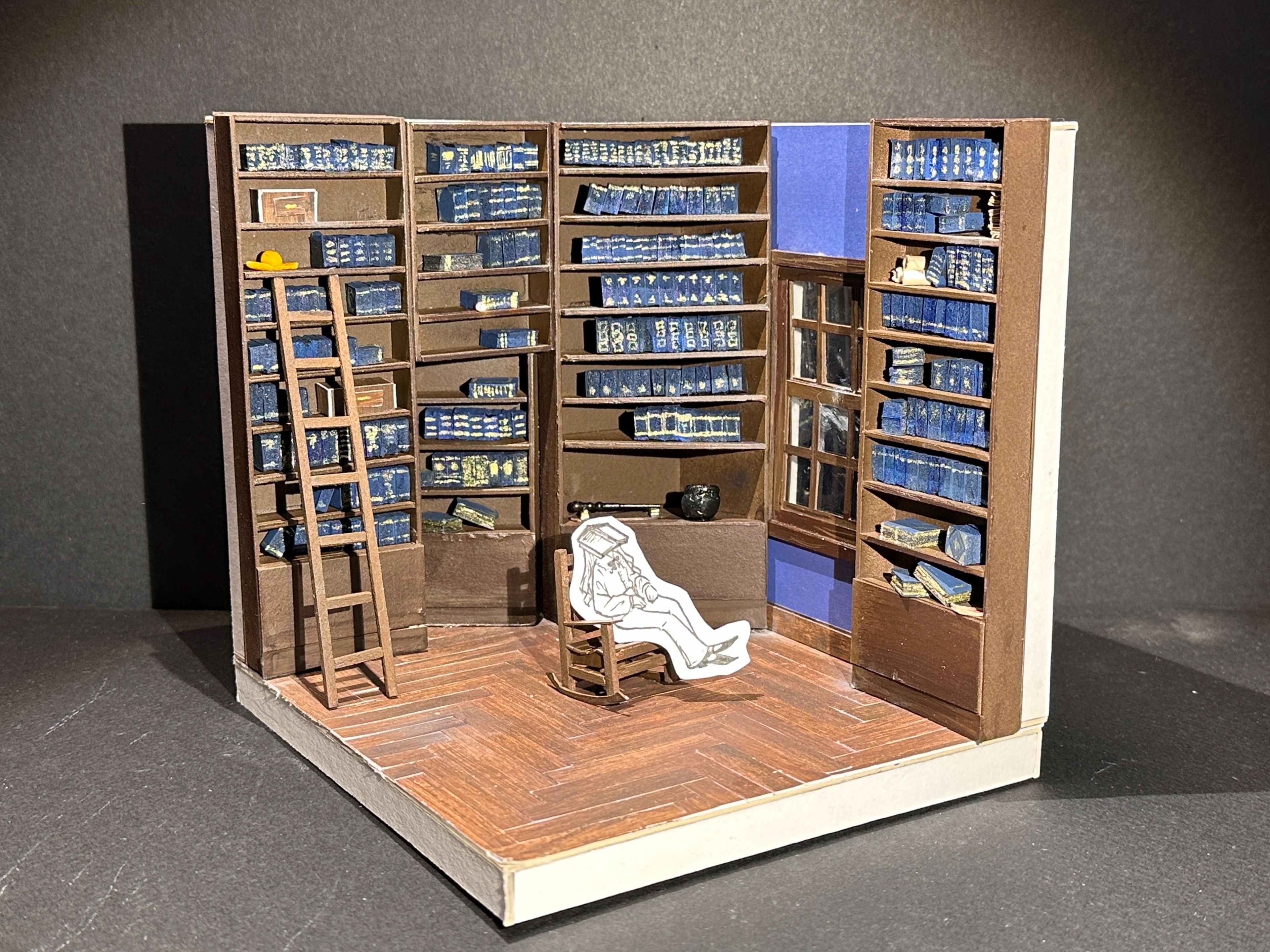 Scale model of a set design for a library, with tall brown bookshelves filled with blue books, a ladder, and a paper cut-out of a person sitting on a chair in the centre.