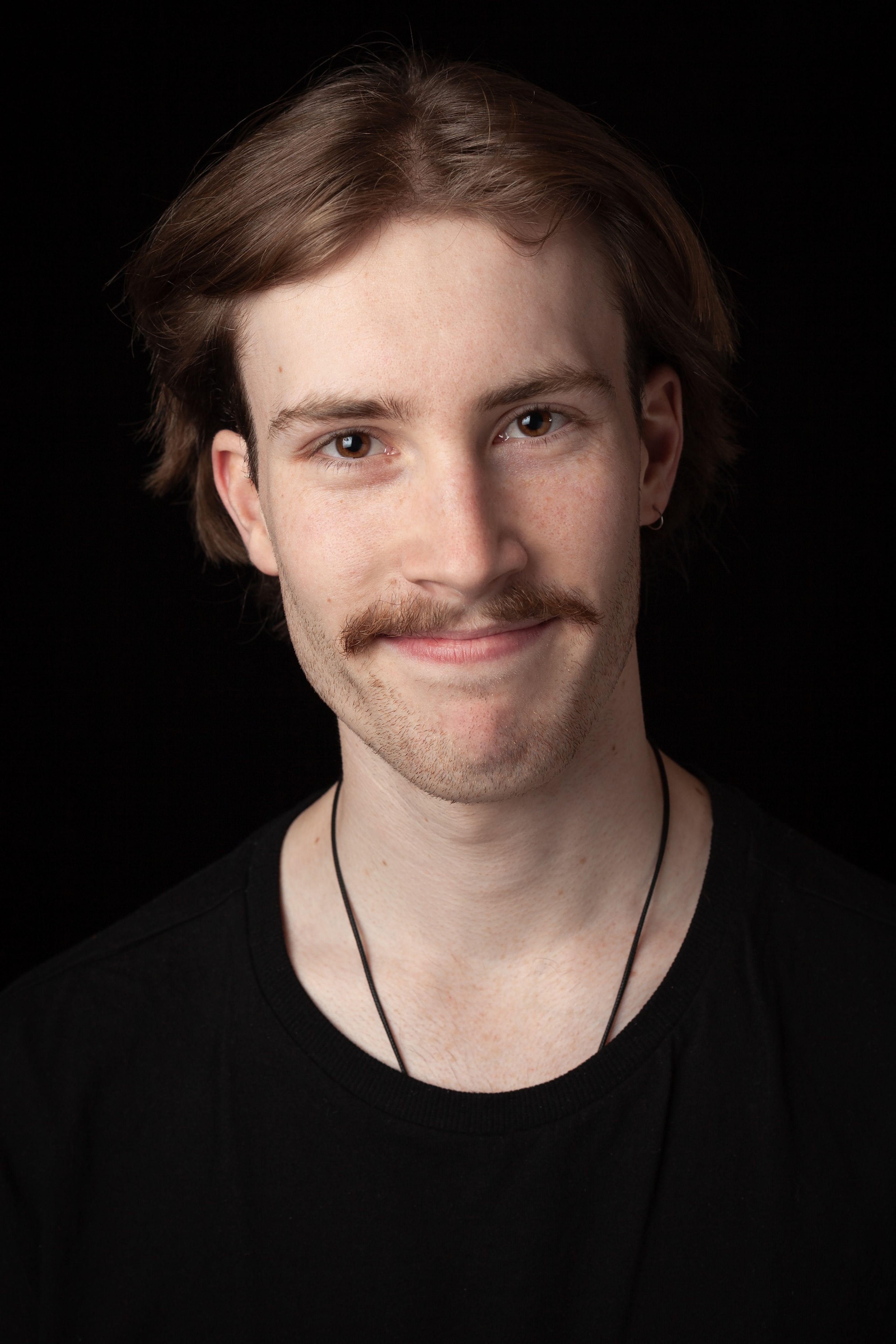 Headshot of a young man with light brown hair and a moustache, wearing a black t-shirt and a simple necklace, against a black background.