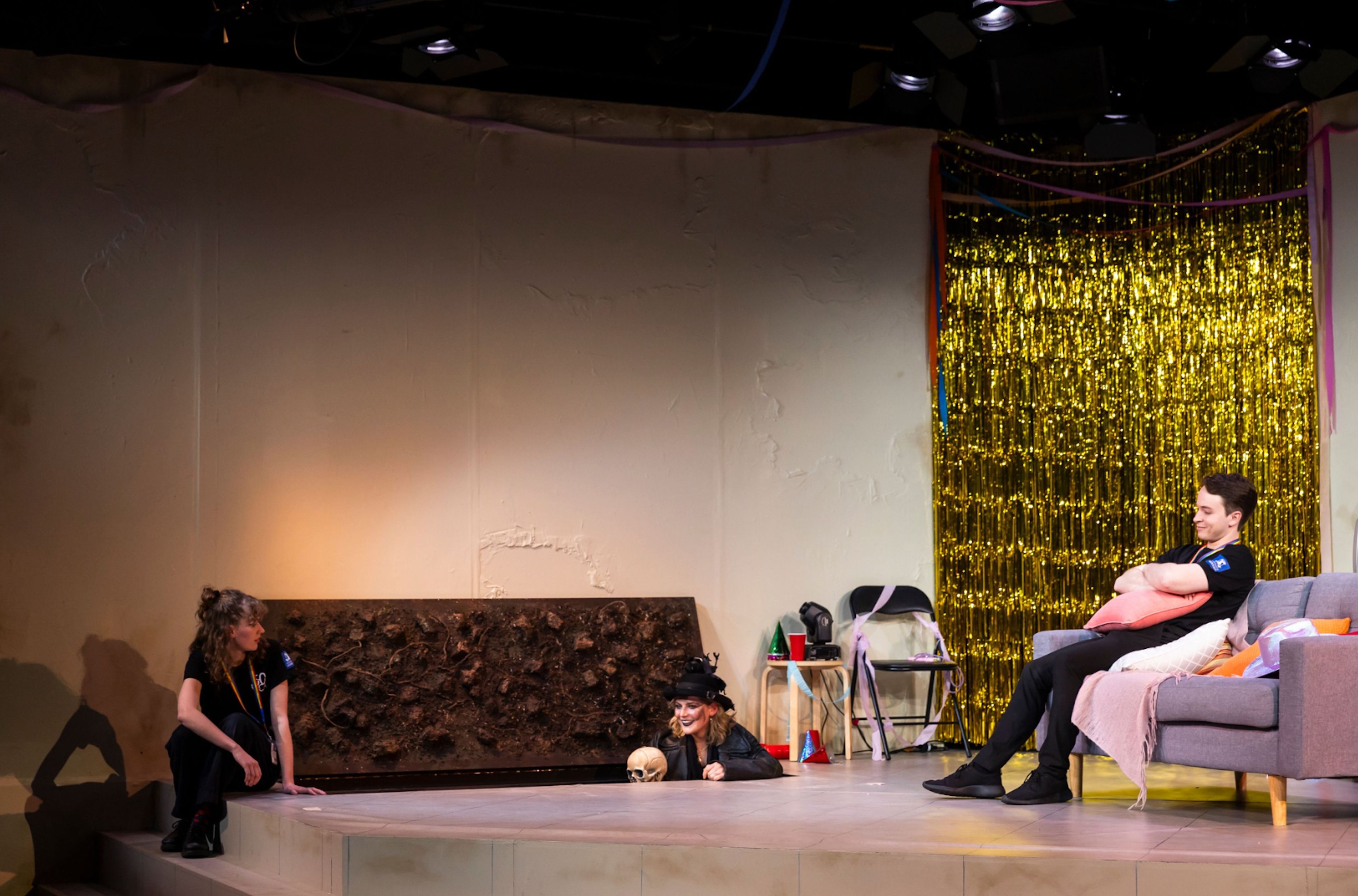 Theatre production photograph showing two actors inside a grave onstage, and another sitting on a couch, with a golden tinsel curtain in the background and festive decorations on the set.