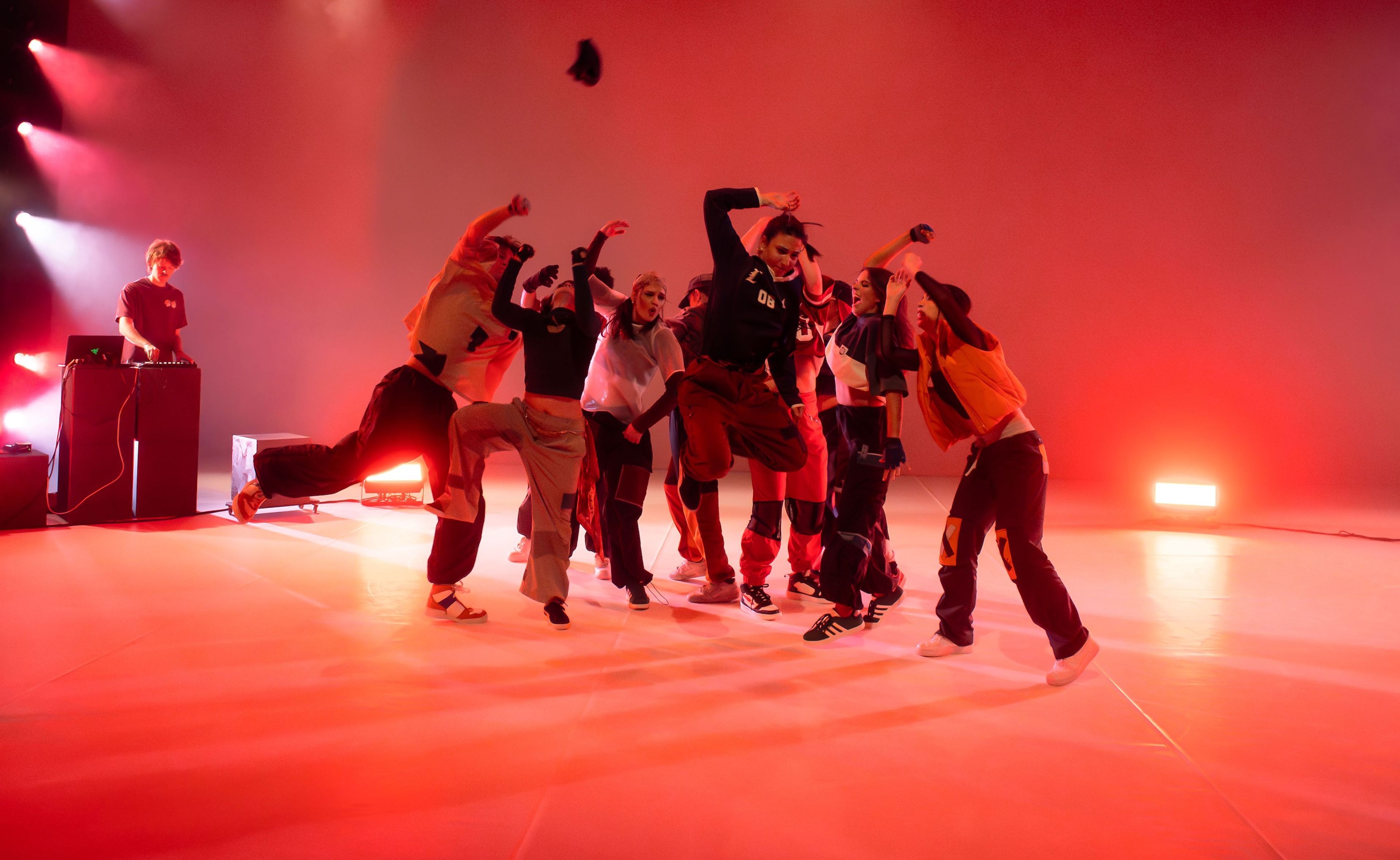 Dance performance photograph with dancers moving dynamically in a warm red-lit environment, with a DJ positioned in the background.